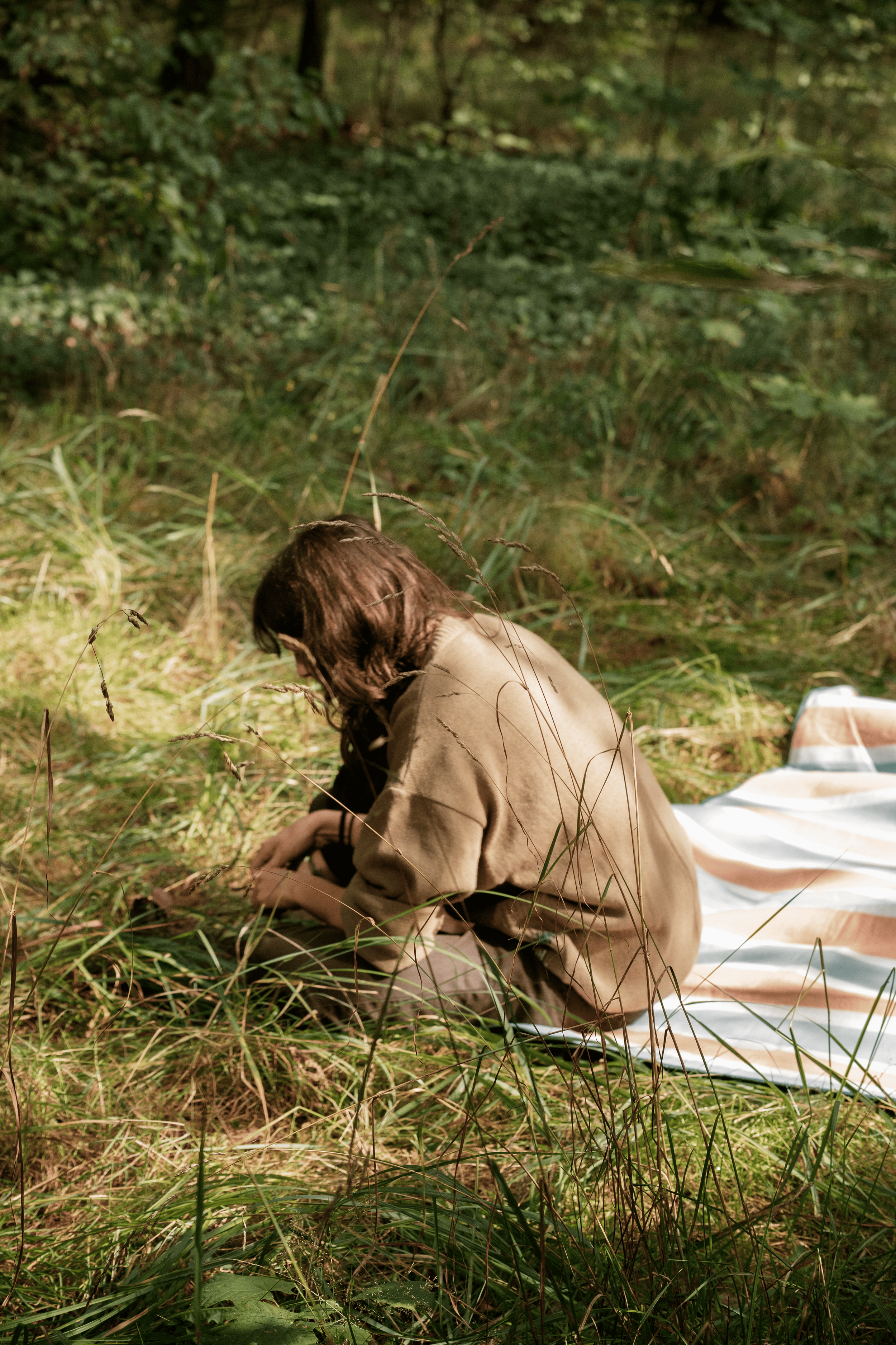 A person with brown hair wearing a beige jacket, sitting on the grass in a wooded area next to a striped blanket.