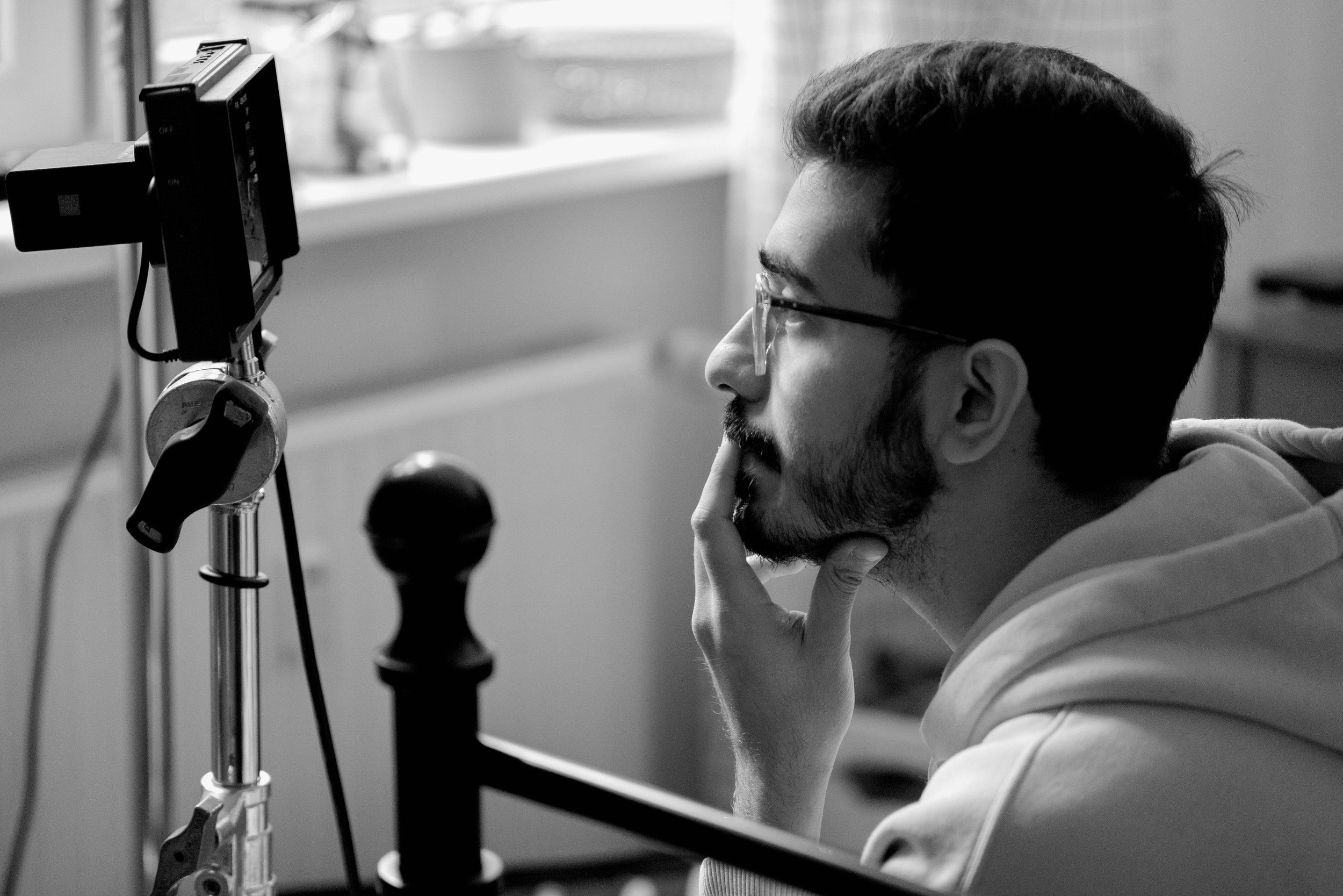 A young man with glasses and a beard sitting at a desk, looking at a computer monitor, with his hand on his chin, in a room with a window.