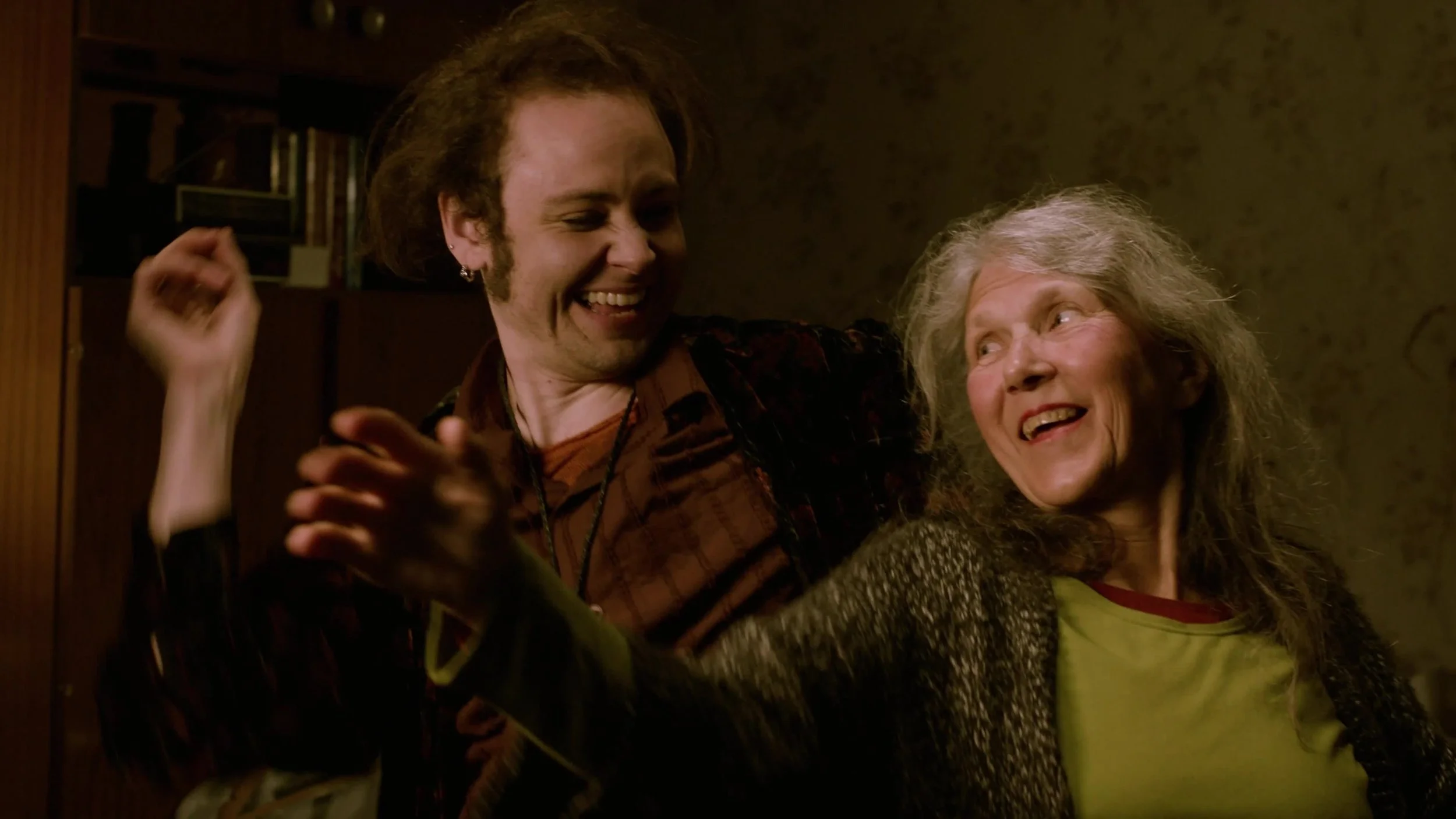 A young man and an elderly woman smiling and dancing together in a cozy, warmly lit room.