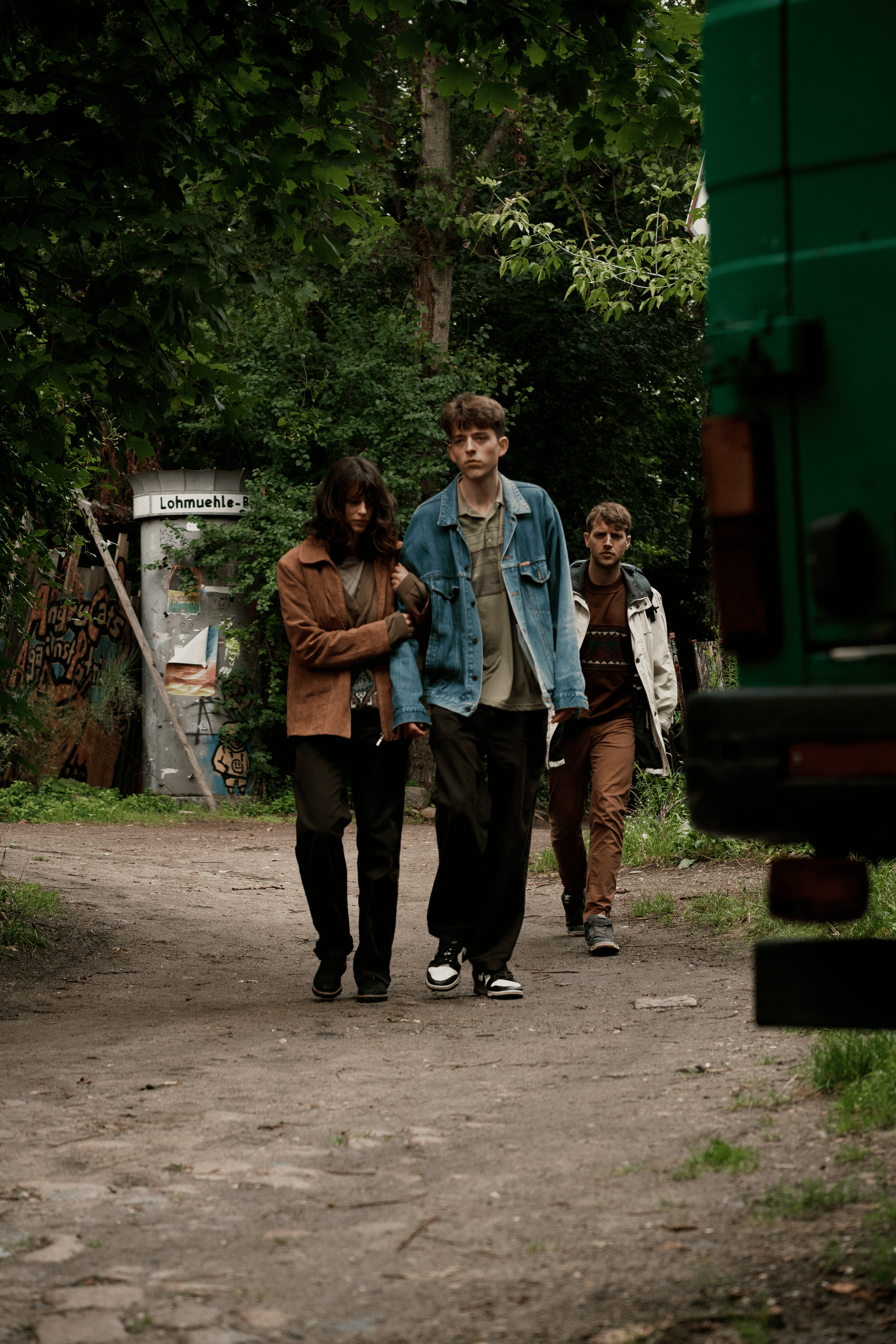 Three young people walking on a dirt path surrounded by trees, with graffiti-covered walls and a green trash bin nearby.