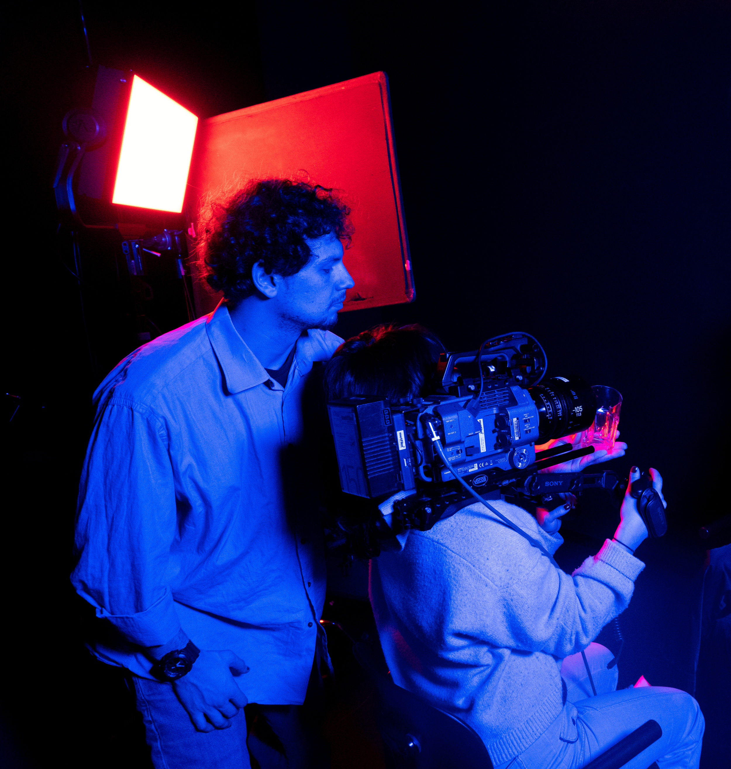 Filmmaker and camera operator working with professional movie camera in a dark studio illuminated by blue and red lighting.