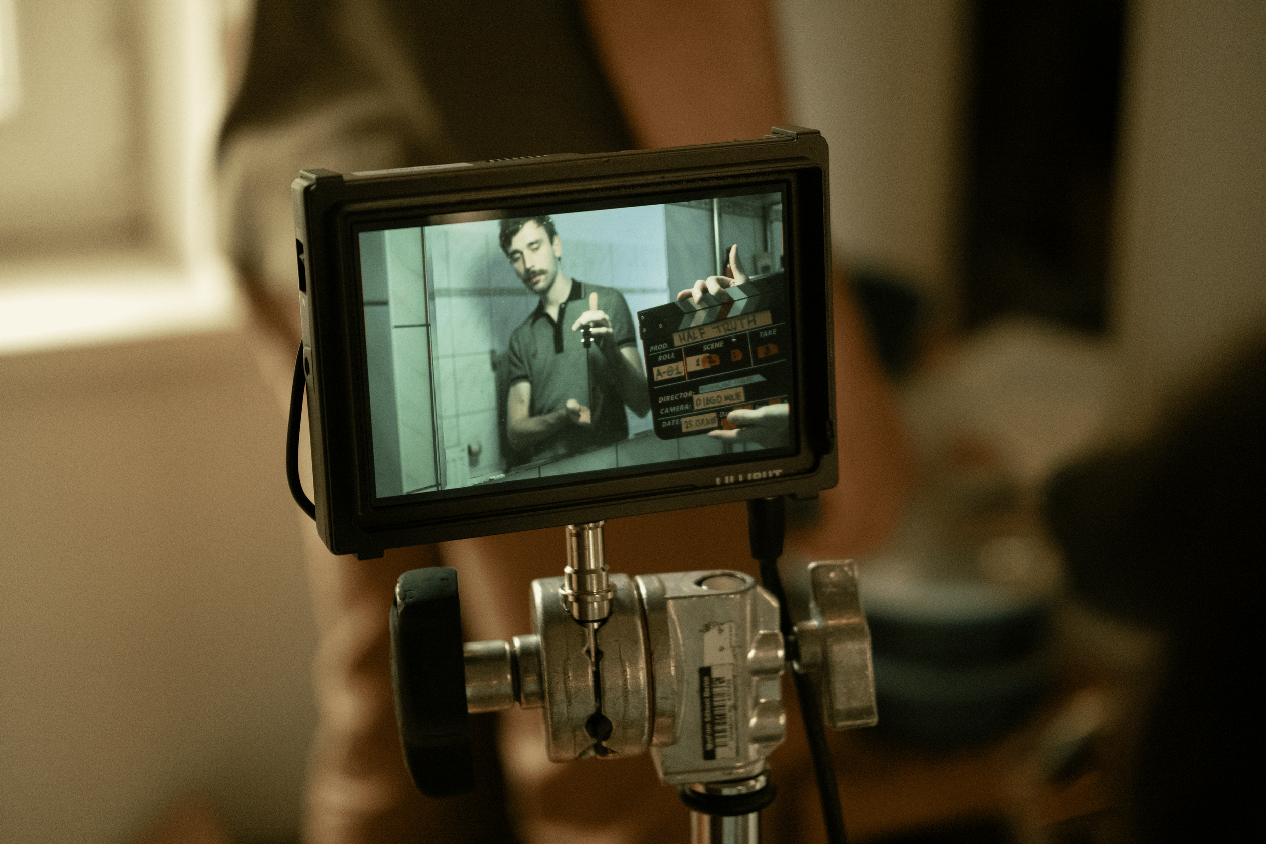 A camera monitor shows a man with dark hair and a beard taking a selfie in a bathroom mirror, making a hand gesture.