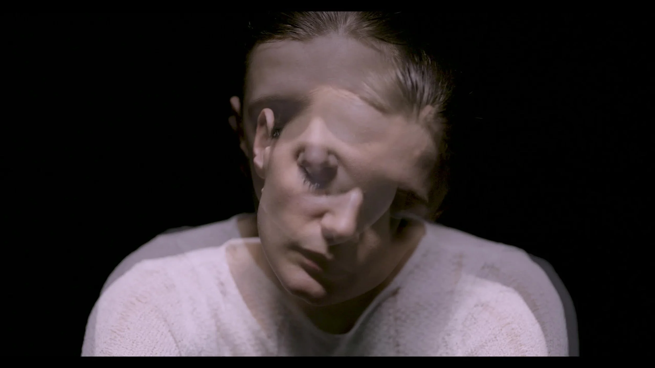 A person with short hair and a light-colored top, blending with a dark background, appears to have multiple overlapping faces, creating a distorted or surreal visual effect.