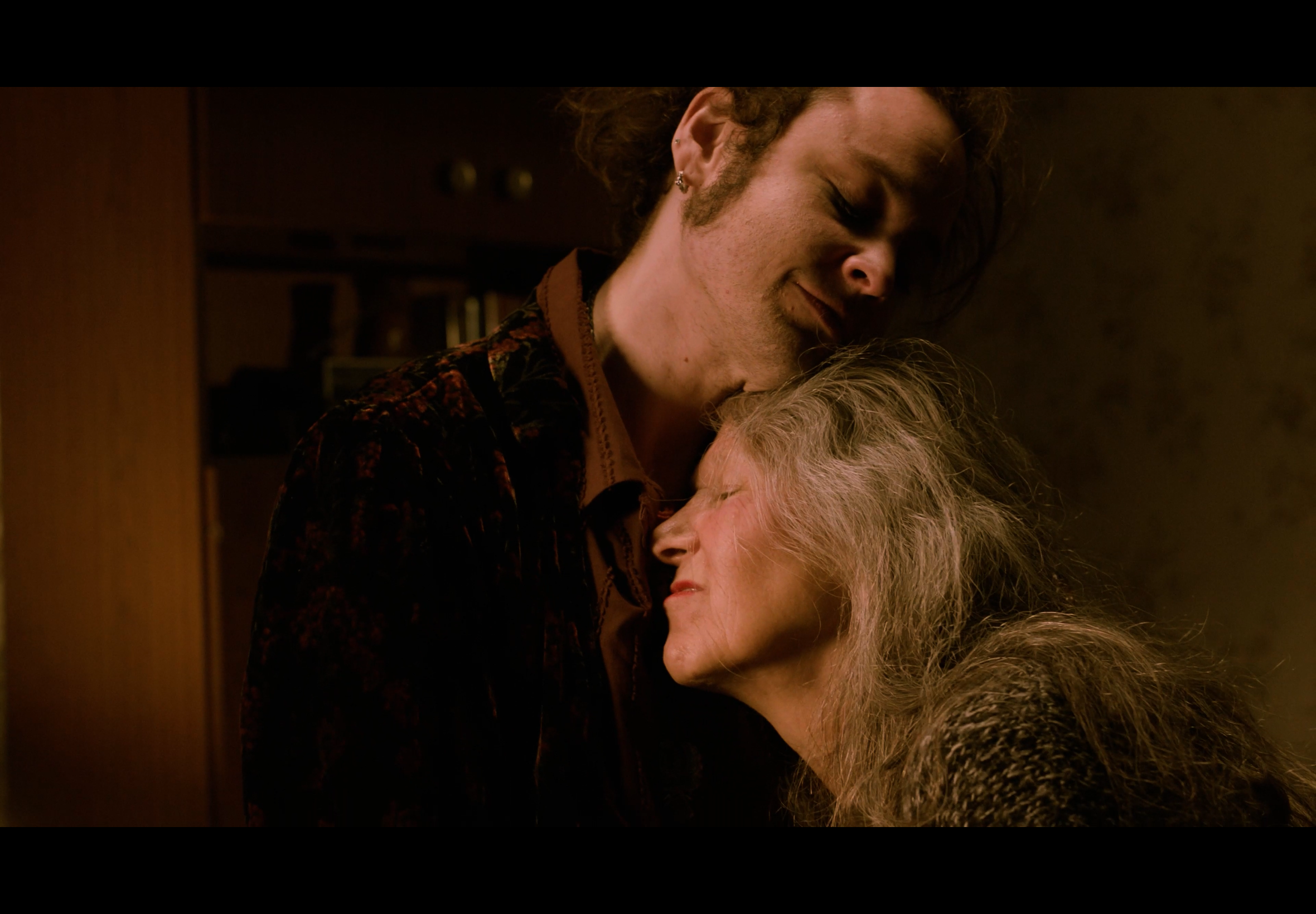 A young man and an elderly woman share a tender moment with their heads touching and eyes closed in a warmly lit room.