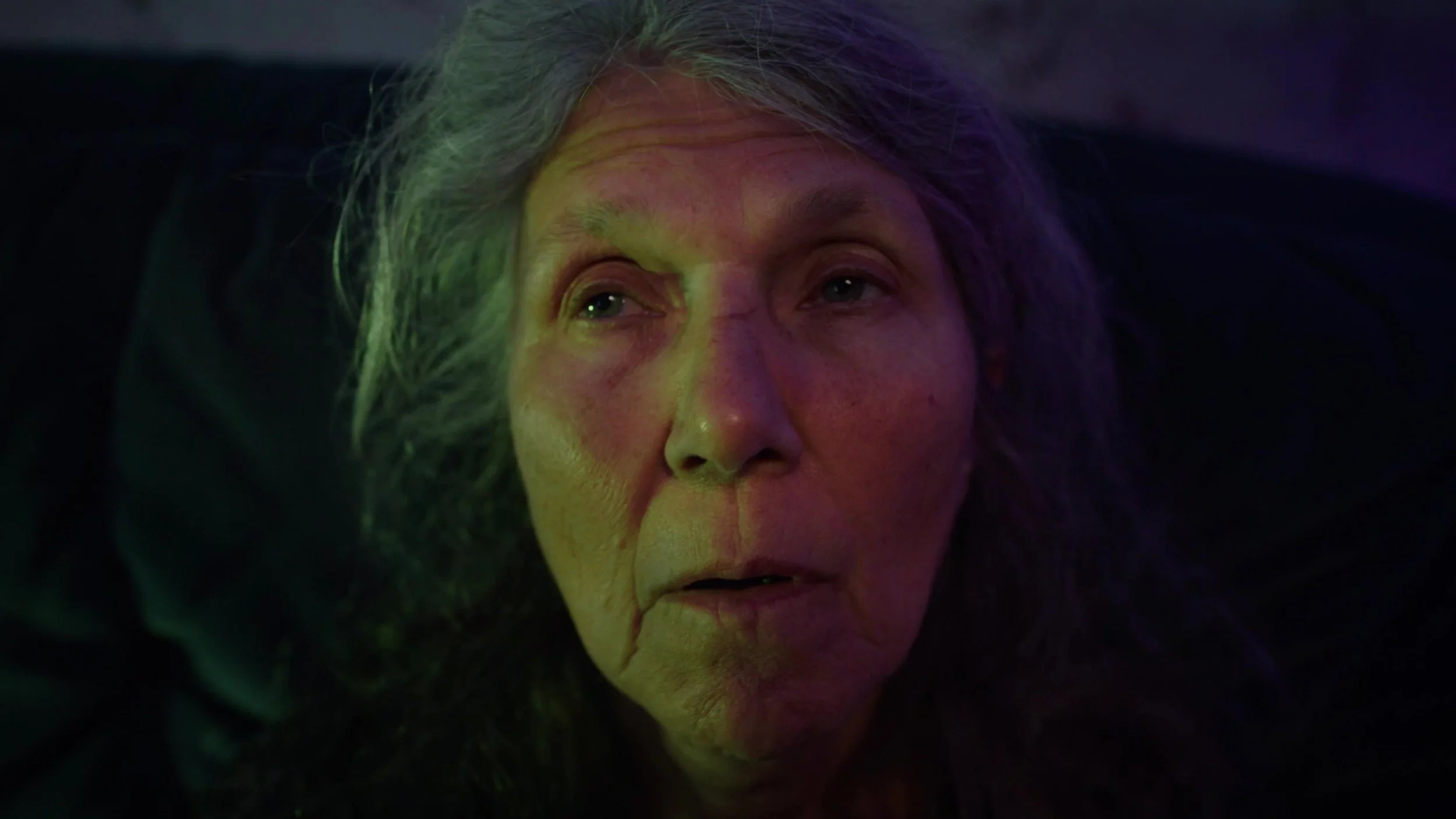 Close-up of an elderly woman with long gray hair, illuminated by colorful lights, with a somber expression.