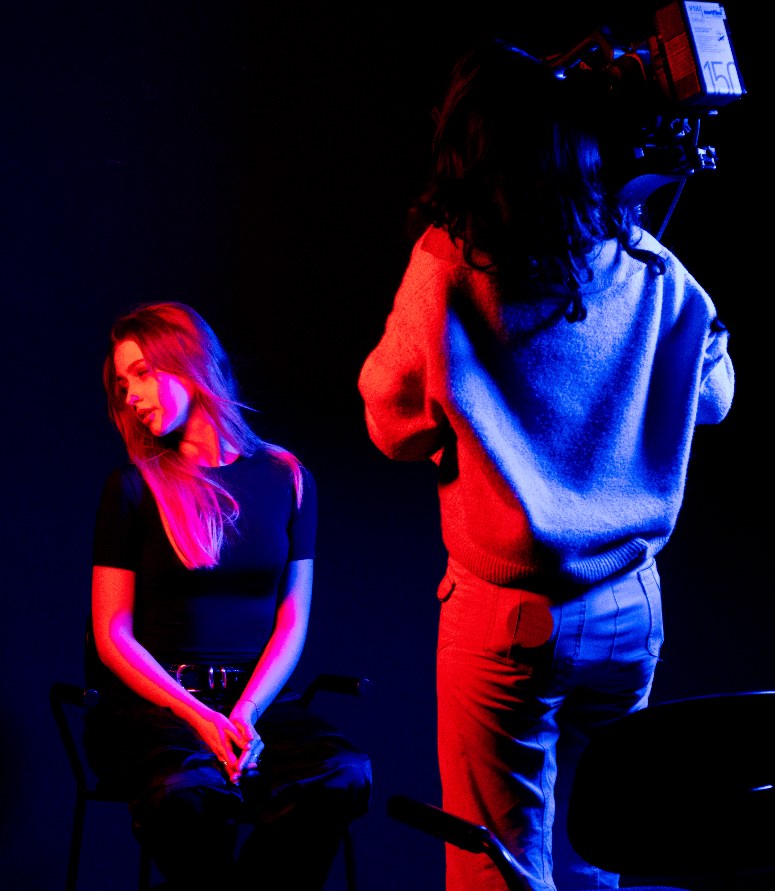 A woman with long blonde hair sitting on a chair during a video shoot, with colorful pink and blue lighting and a crew member operating a camera.