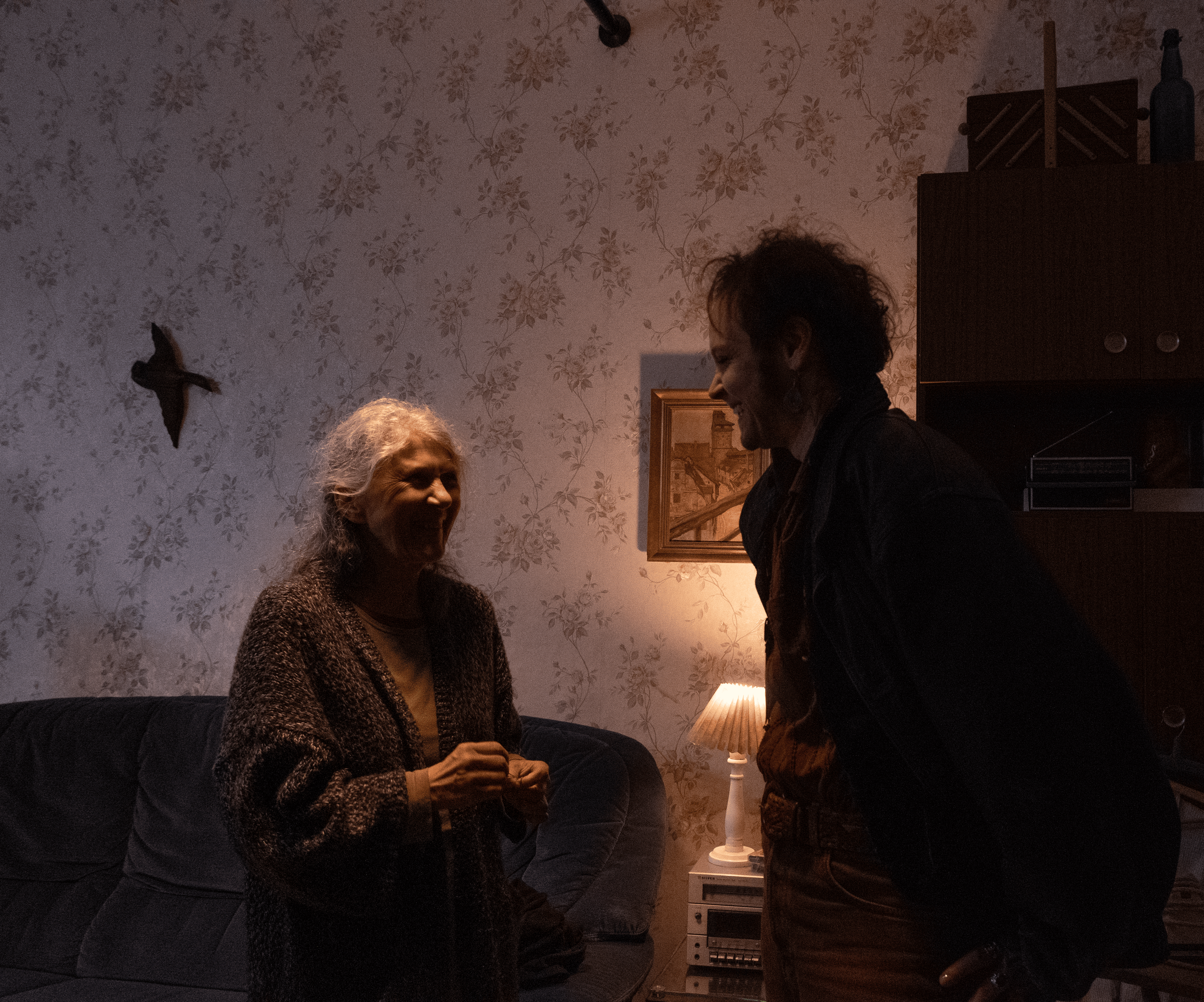 A woman and a man are happily conversing inside a living room, with dim lighting from a table lamp, wallpaper with floral pattern, and wall art in the background.