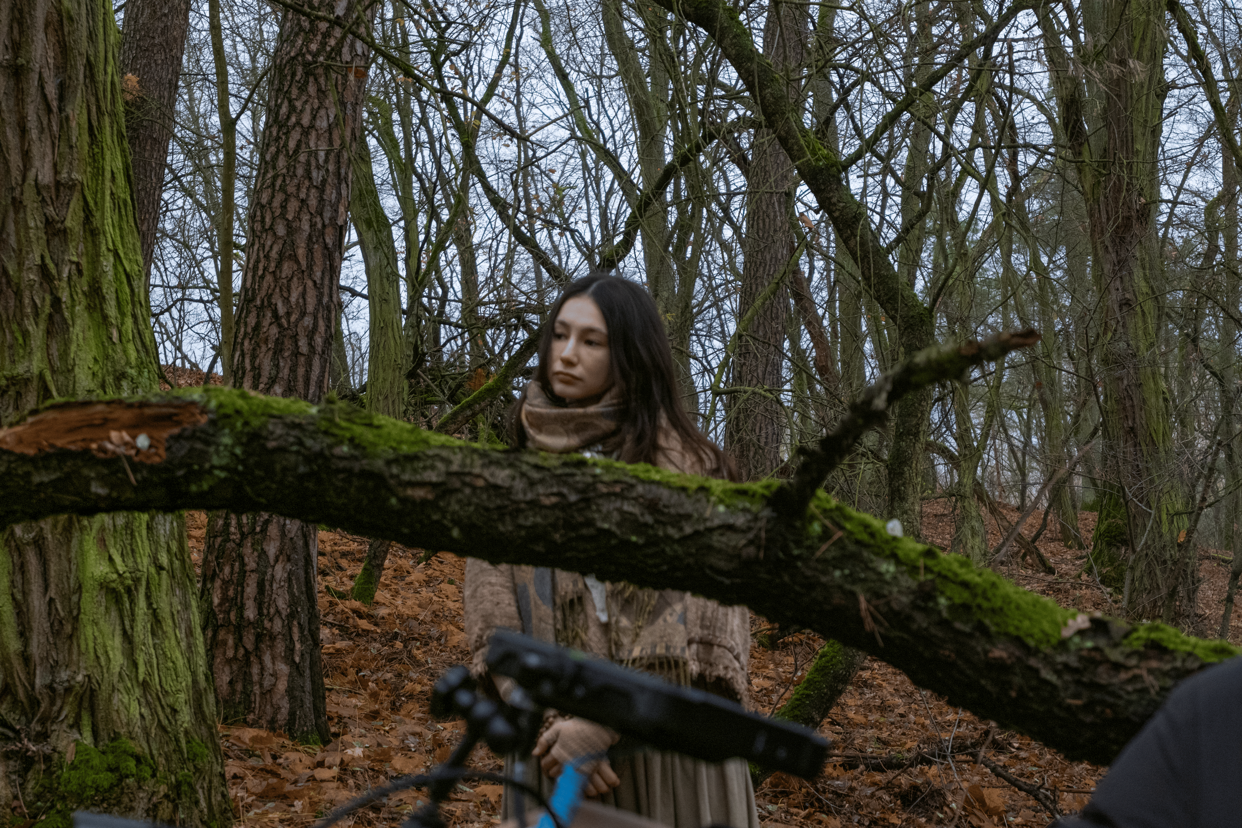 A woman with long dark hair wearing a beige coat and scarf standing in a wooded area with leafless trees and fallen leaves on the ground, looking to the side.