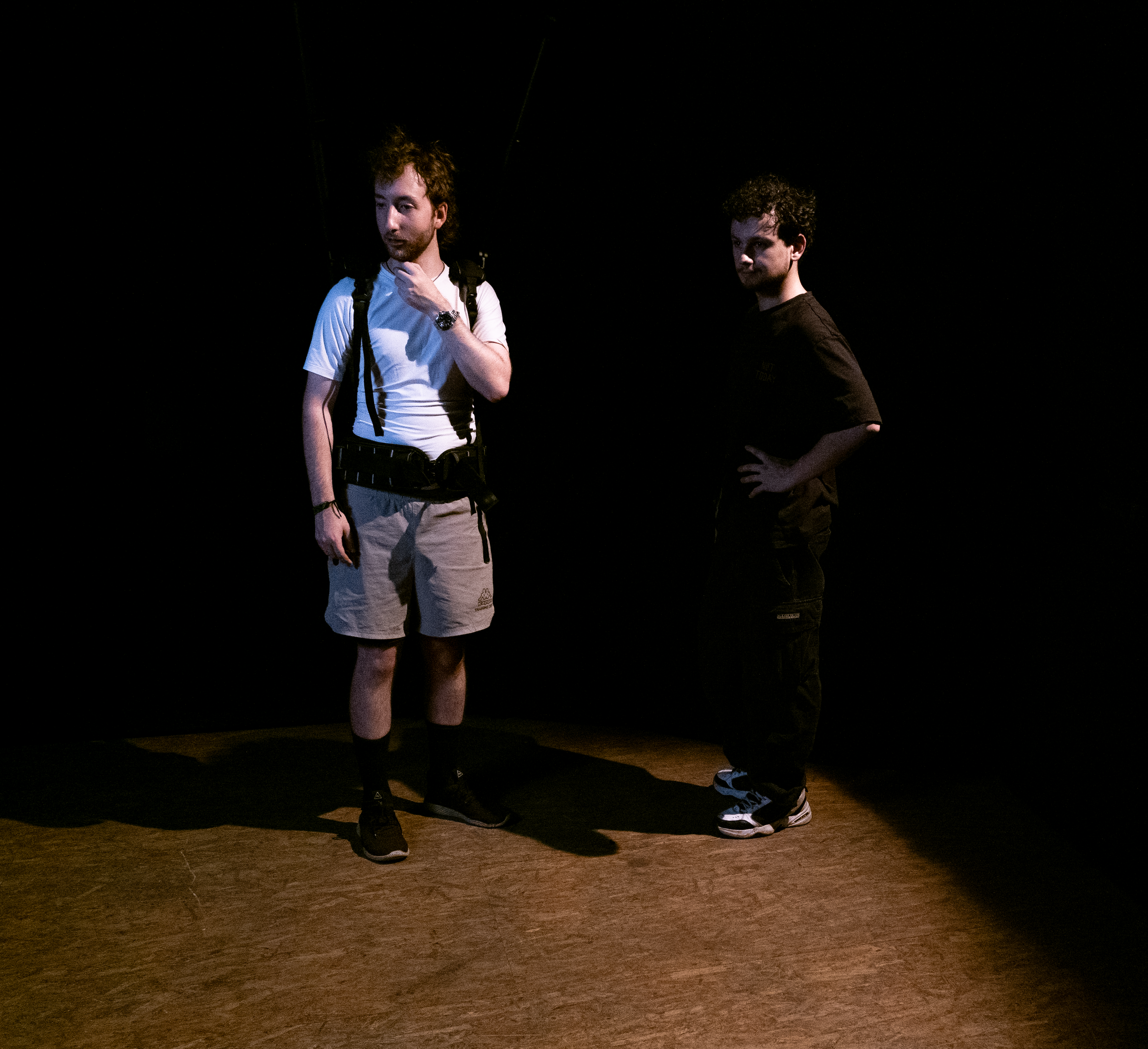Two young men standing in a dark room with a illuminated floor. The man on the left has curly hair, a beard, and is wearing a white t-shirt, beige shorts, and black socks with sneakers. He is touching his chin with his right hand and has a black back