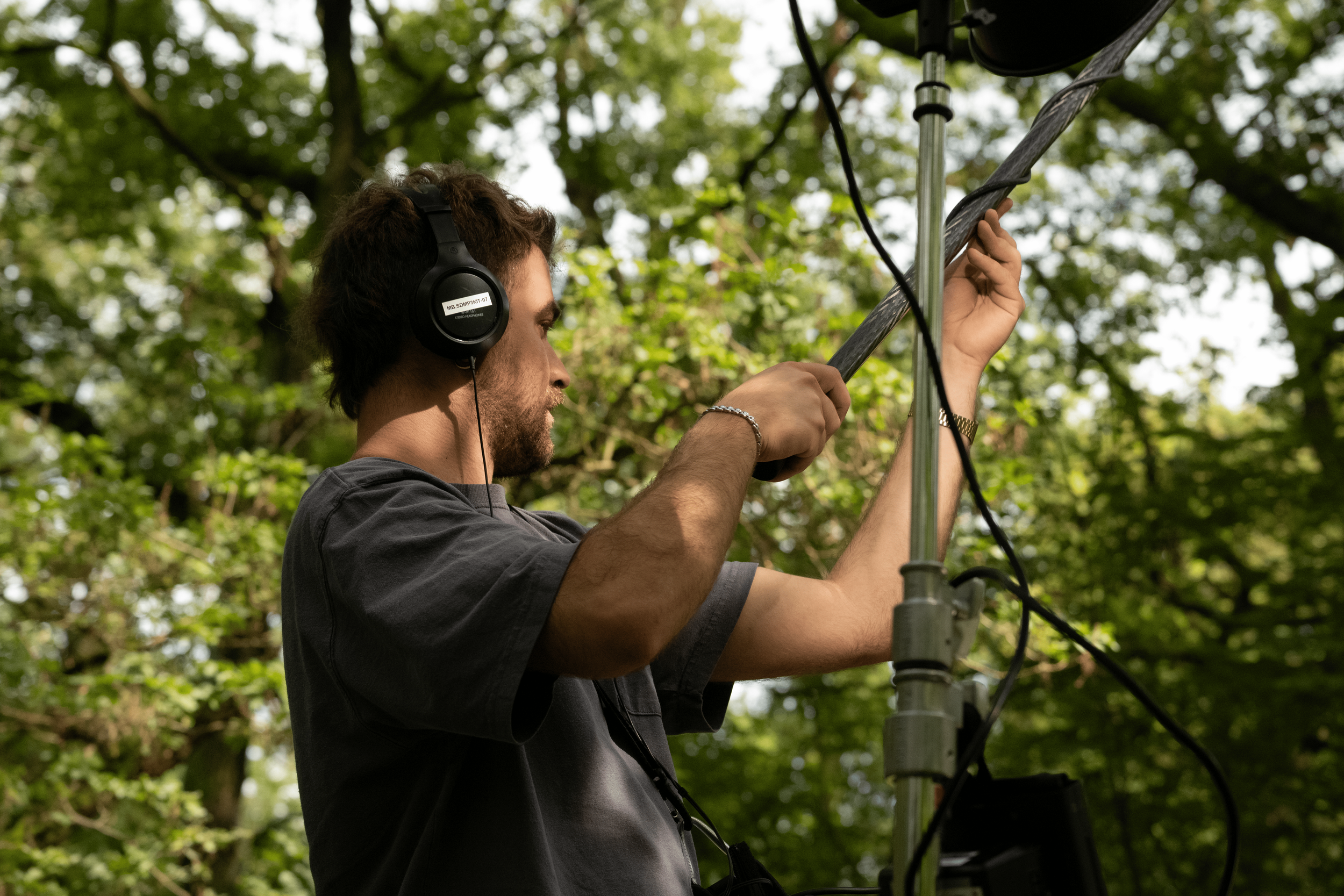 A man wearing headphones adjusts equipment outdoors amidst green trees.