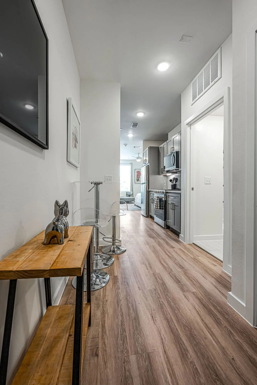 Interior view of an apartment with a hallway leading to a kitchen and living area, wooden floor, with a wooden console table and decorative dog sculpture on the left, and a wall-mounted tv on the left wall.