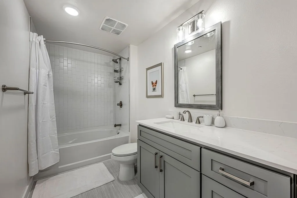 A modern bathroom with a bathtub and shower, white tile surround, a white toilet, a gray vanity with a white marble countertop, a framed picture of a fox, and a mirror with overhead lighting.