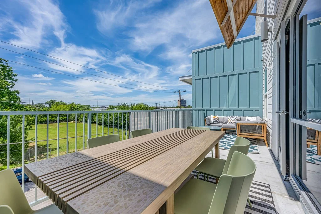 Balcony with outdoor furniture, including a large dining table with chairs and a sitting area with a sofa and chairs, overlooking a green field and blue sky with clouds.