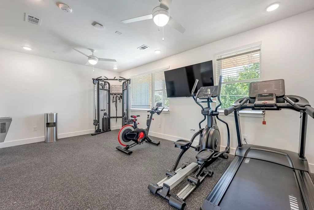 Home gym with treadmill, elliptical, stationary bike, weight machine, a large wall-mounted TV, windows with blinds, and ceiling fans.