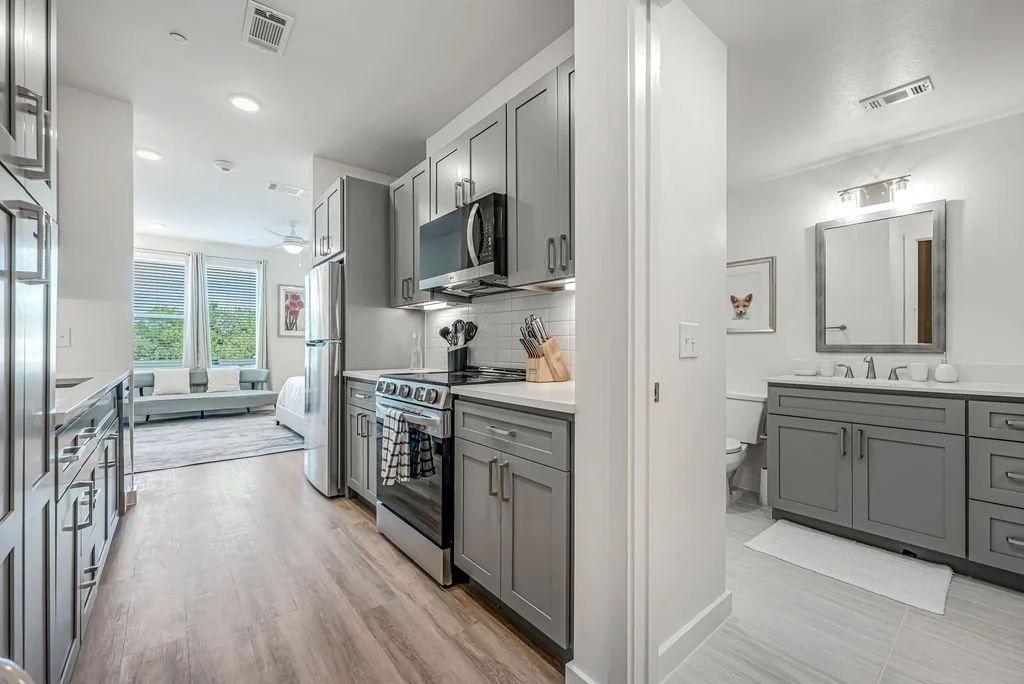 Open-concept kitchen with gray cabinets, stainless steel appliances, and a view into a bright living room with large windows and a white sofa.