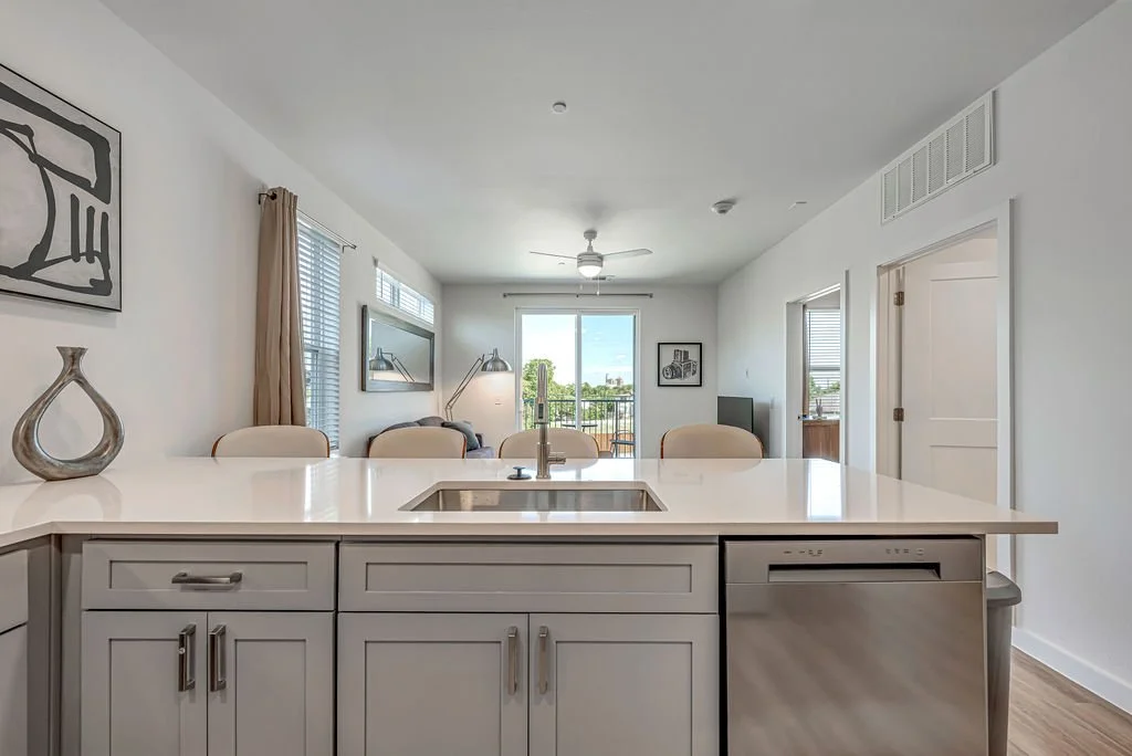 Modern open-concept living room and kitchen with white cabinets, a kitchen island with a sink, and a view of a balcony with sliding glass doors.