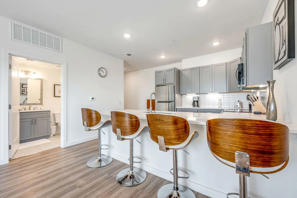 A modern kitchen with gray cabinets, white countertops, and four wooden barstools at the island, with a view into a bathroom.