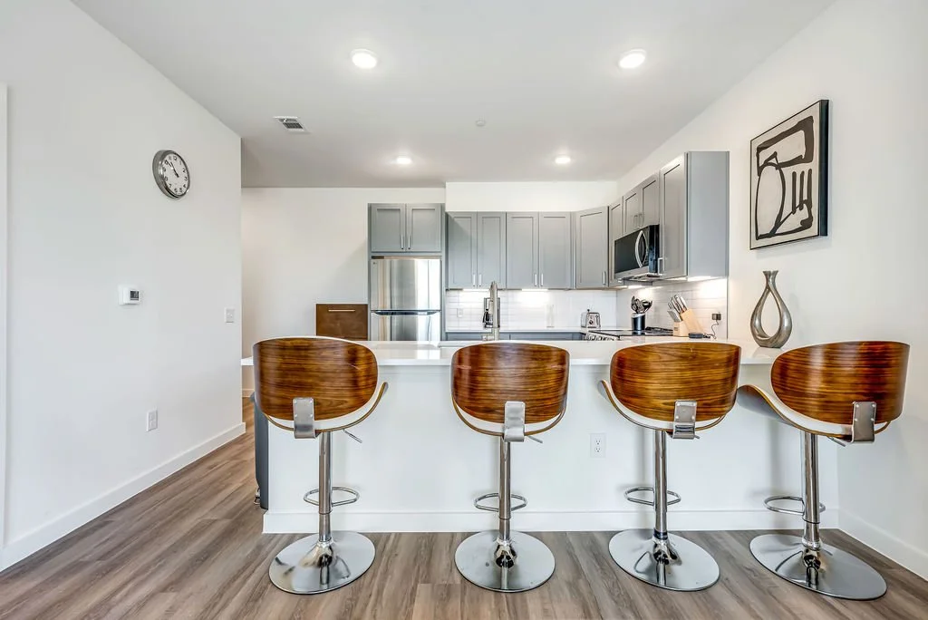Modern kitchen with four wooden bar stools at a white counter, gray cabinets, stainless steel appliances, and minimalist wall art.