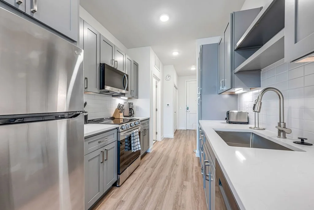 Modern kitchen with gray cabinets, stainless steel appliances, white countertops, and light wood flooring.