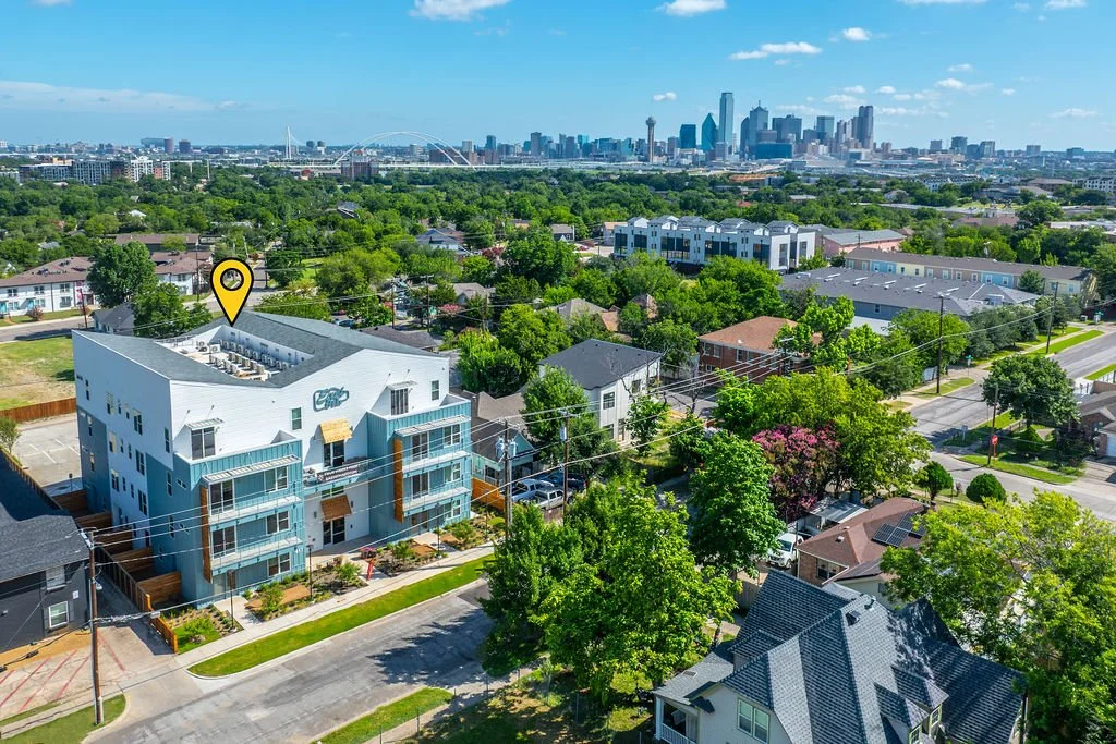 Aerial view of a residential neighborhood with a building marked with a yellow location pin, overlooking the Dallas skyline in the background, with lush green trees and streets in the foreground.