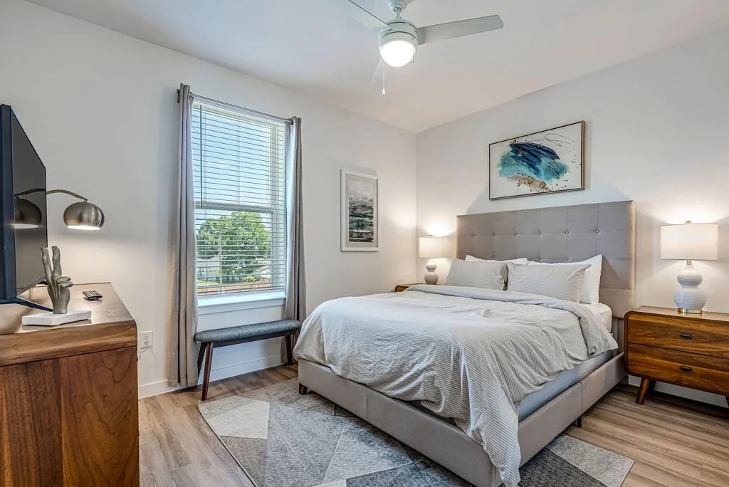 A bedroom with a large bed, two nightstands with lamps, a window with blinds, and a ceiling fan.