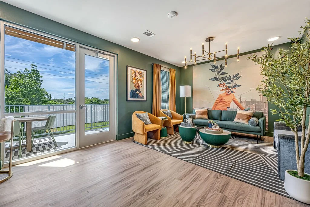 Bright living room with large window and glass door leading to balcony. Green walls, modern chandelier, artwork, yellow chairs, and a teal sofa with decorative pillows. Potted plant and a black-and-white striped rug.