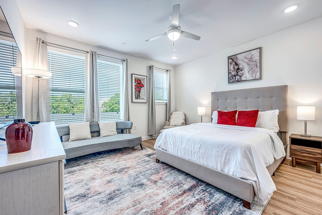 A modern bedroom with a large bed, two nightstands with lamps, a gray upholstered headboard, a ceiling fan, and a sitting area with a gray sofa and pillows near windows with white blinds and curtains.