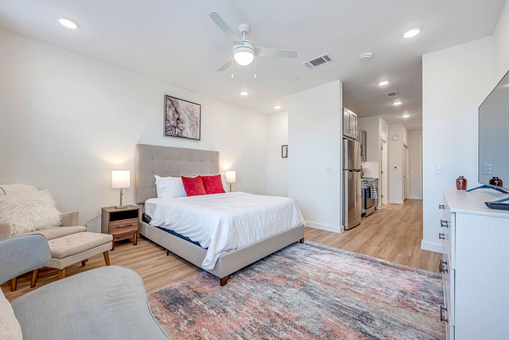 A modern studio apartment featuring a bed with a tufted beige headboard, two red pillows, and white bedding. Beside the bed are two matching nightstands with lamps. There's a beige armchair with a fluffy pillow, a large rug, and a white dresser with 