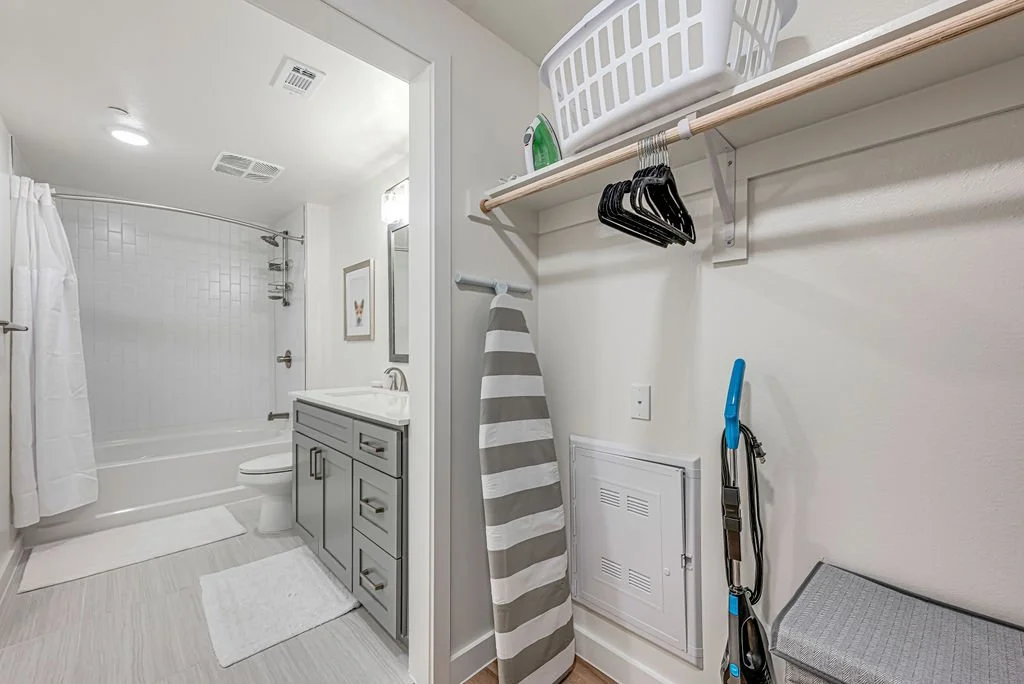 Bathroom with white tiles, shower with white curtain, gray vanity with sink, and small framed art on the wall. Closet area with a shelf holding a laundry basket, iron, and a hanging bar with empty hangers. An ironing board is leaned against the wall,