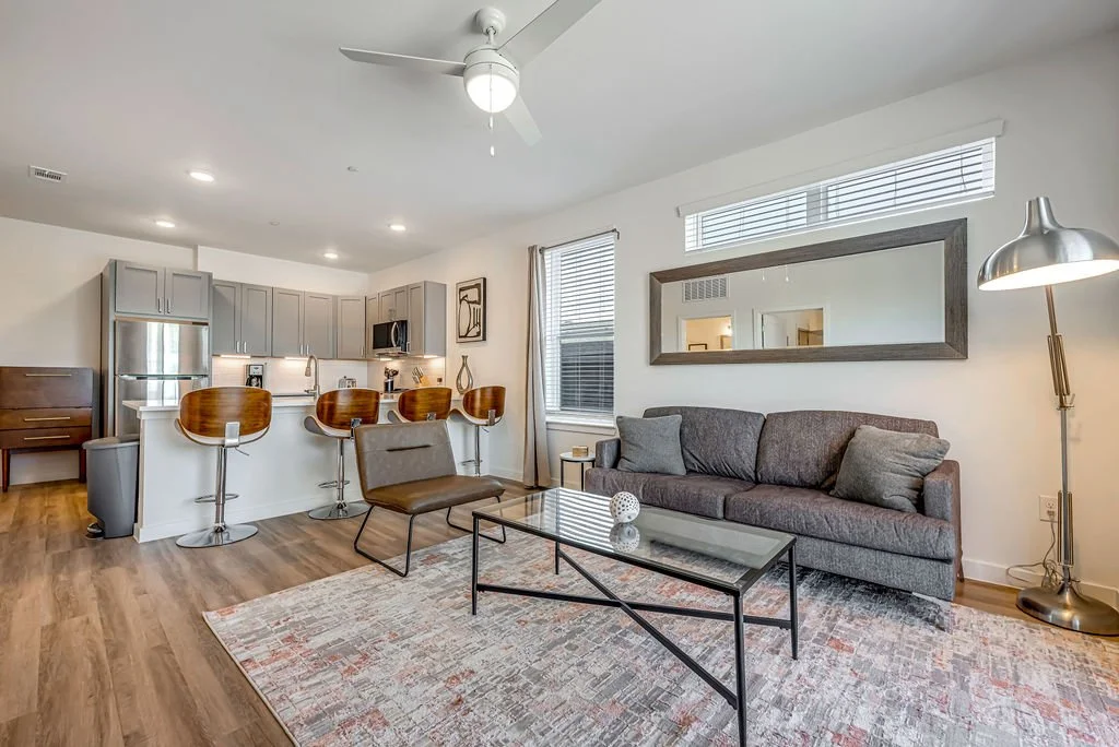 Living room with a gray couch, glass coffee table, and a tall silver floor lamp, open to a kitchen with gray cabinets, a white countertop, and four wooden barstools.