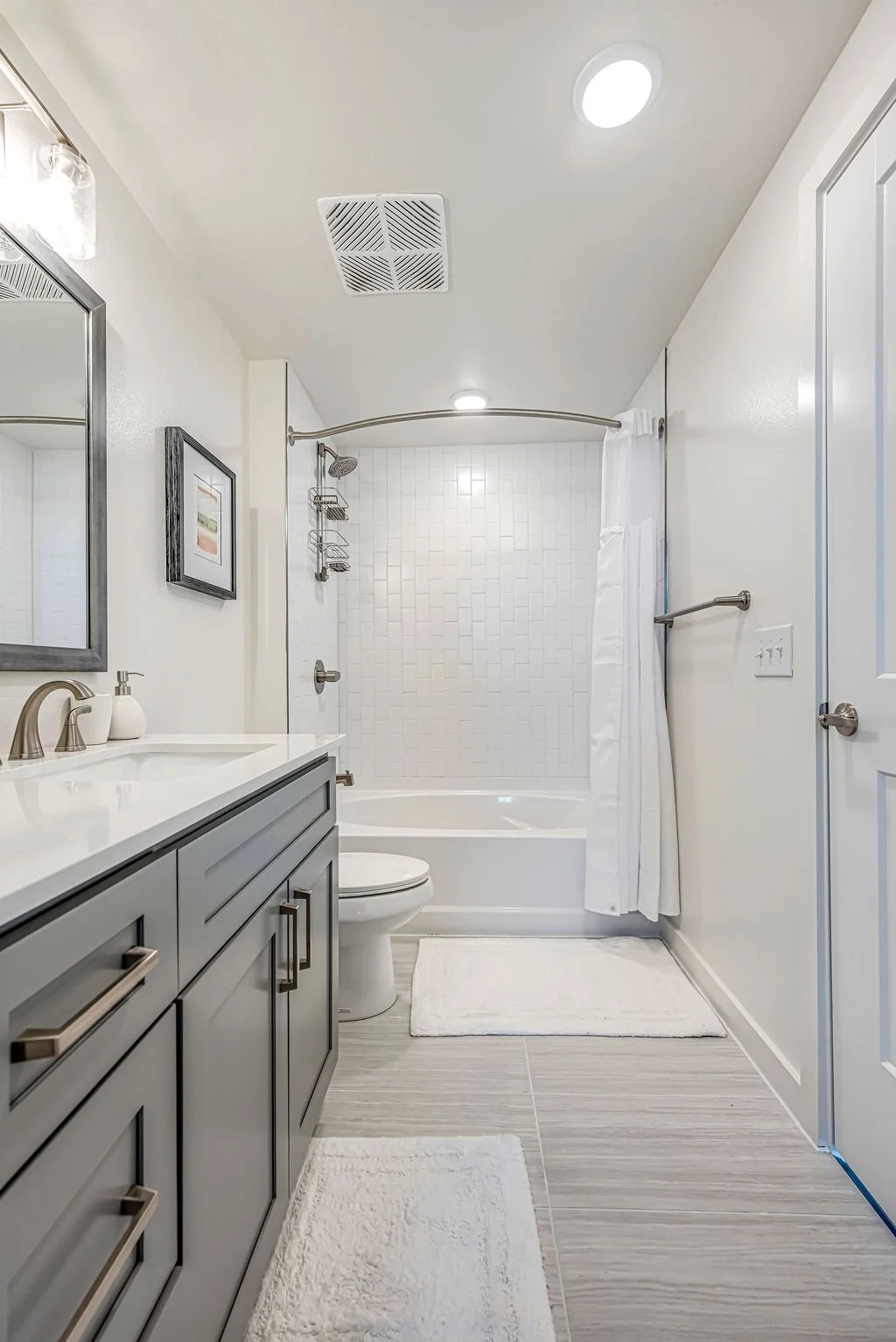 Modern bathroom with gray cabinetry, white countertop, white toilet, bathtub with shower curtain, white tiled shower wall, and bathroom rugs.