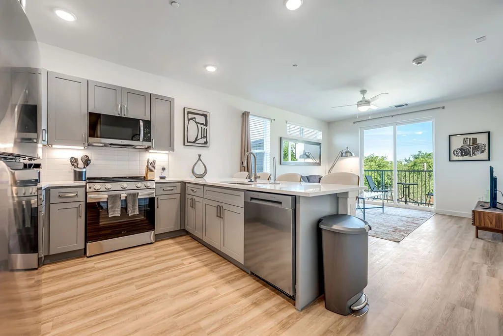 Open-concept kitchen and living room with gray cabinets, stainless steel appliances, and hardwood flooring, leading to a balcony with outdoor seating.