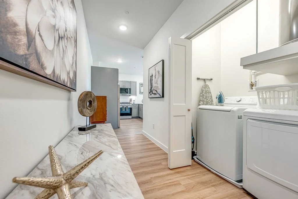 Laundry room with washer and dryer, ironing board, and laundry basket, open to a hallway leading to a kitchen.