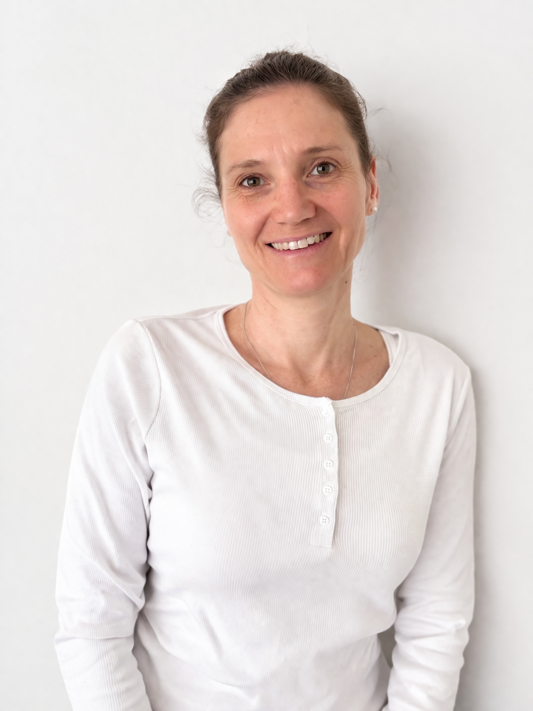 Lächelnde Frau mit braunen Haaren in einem weißen Hemd, vor weißer Wand.