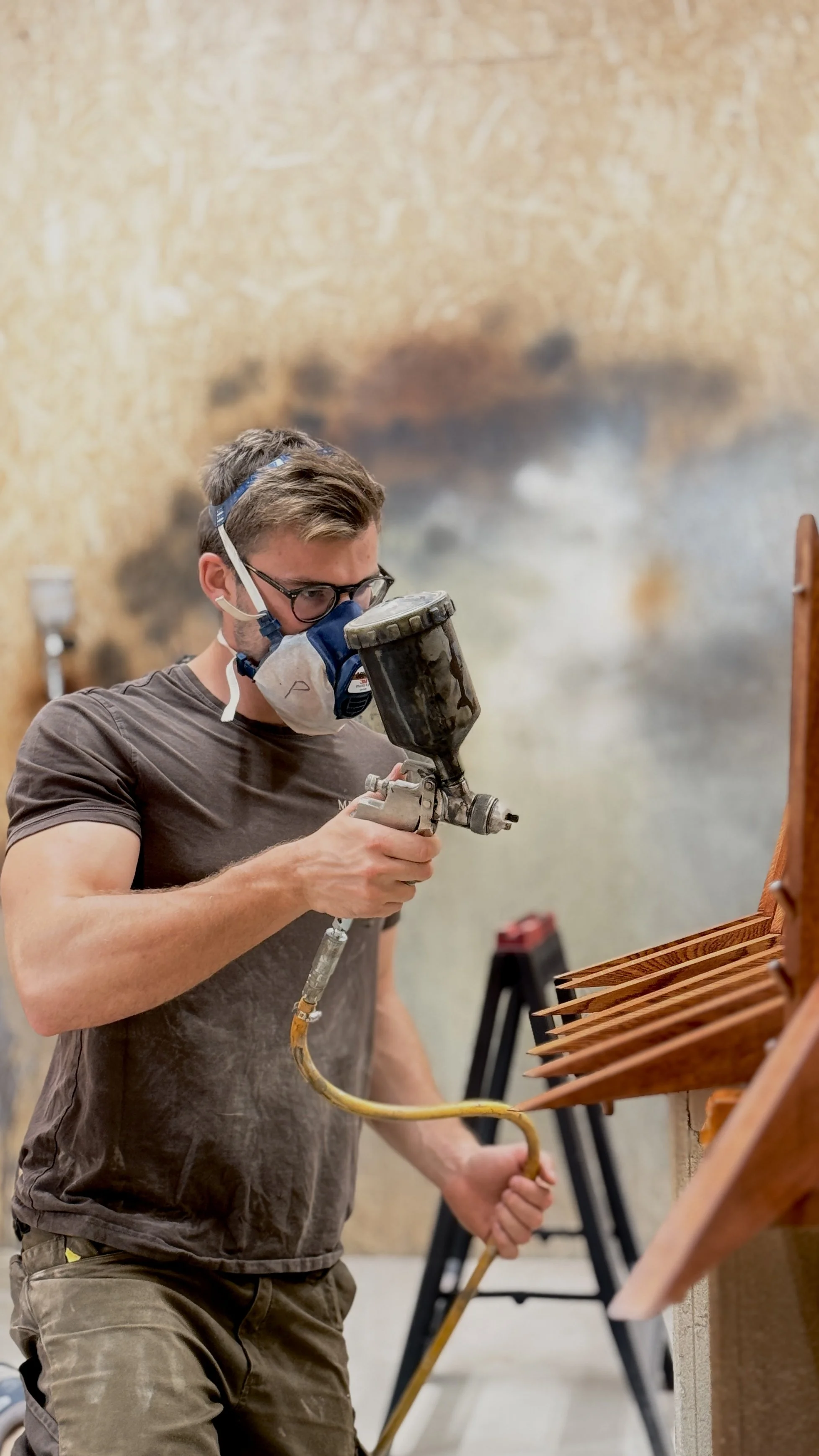 Person spray painting a wooden table with a spray gun, wearing protective gear.