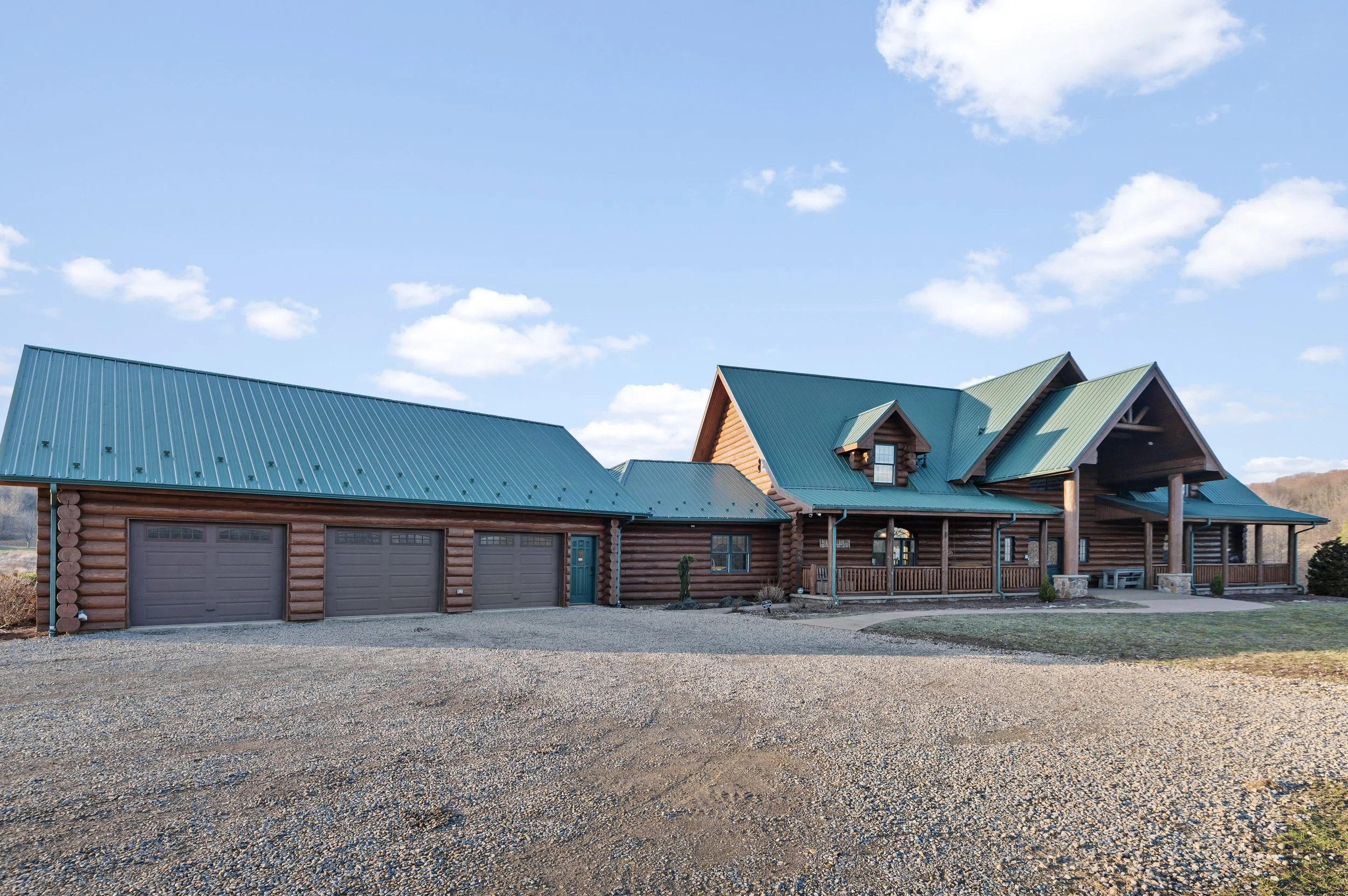 A large log cabin with a green metal roof, three garage doors, and a spacious front porch, set against a blue sky with scattered clouds.