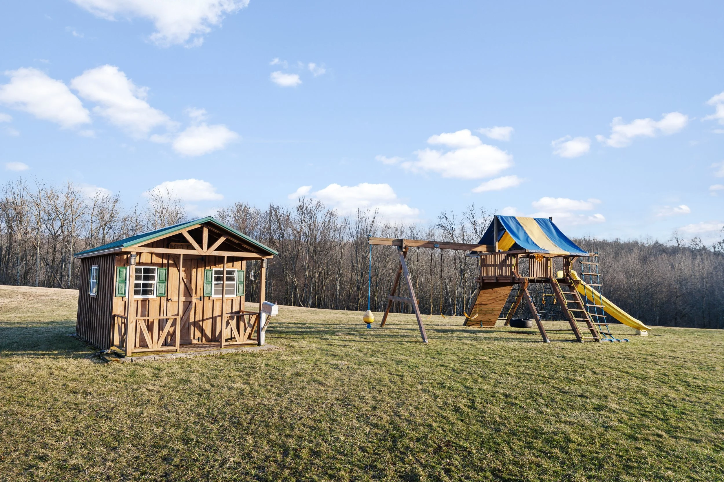 A backyard with a wooden playhouse, a swing set with a yellow slide and a blue and yellow canopy, and a grassy field with leafless trees in the background under a blue sky with scattered clouds.