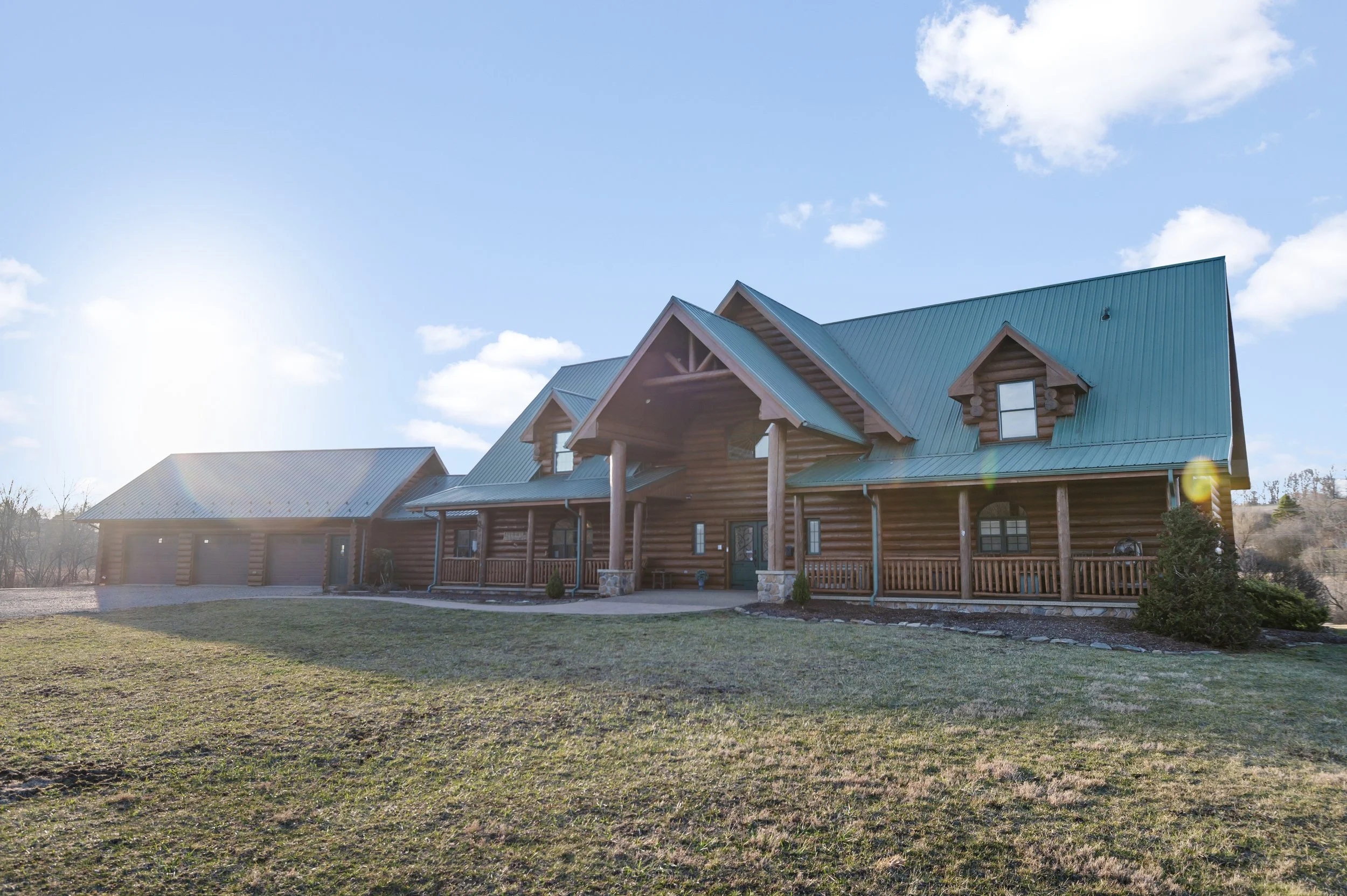 A large log cabin with a teal metal roof, front porch, and attached garage, surrounded by a grassy yard with a few small bushes, under a bright blue sky with some clouds.