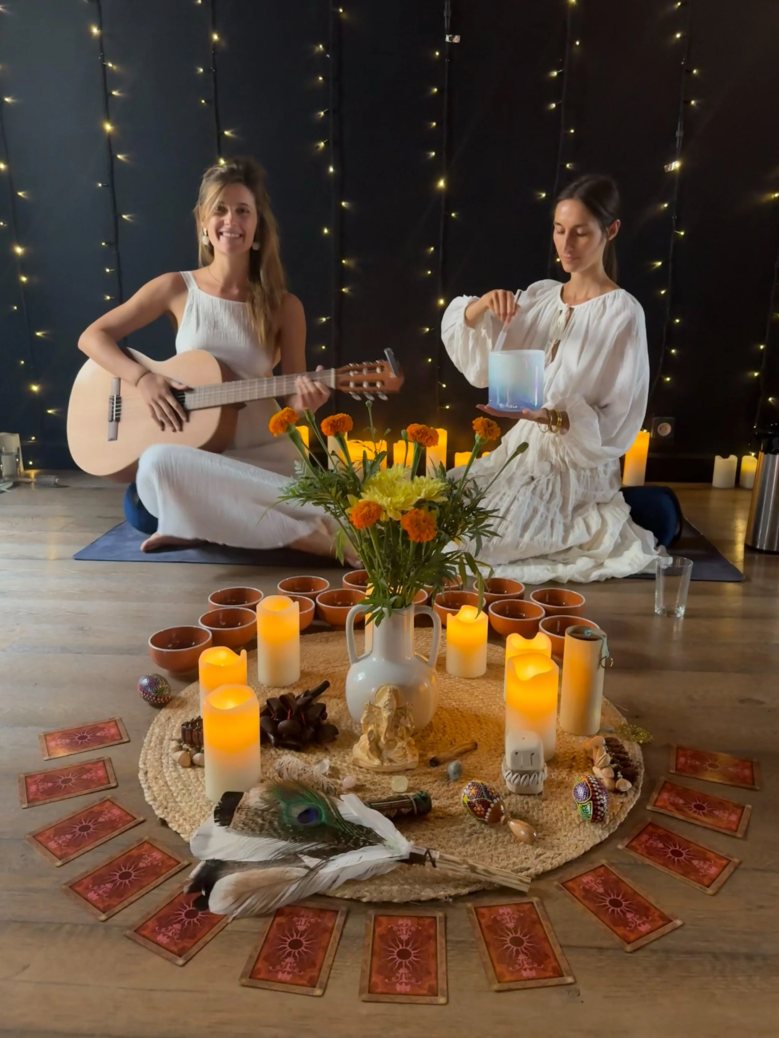 Deux femmes, l'une jouant de la guitare et l'autre tenant un bol, sont assises en méditation dans une ambiance chaleureuse avec bougies, fleurs, cartes de tarot, et décorations spirituelles.