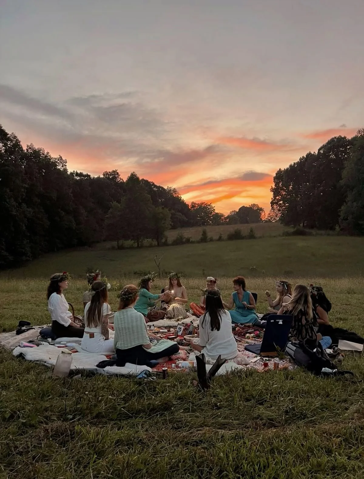 Groupe de personnes assises en cercle sur une prairie lors d'un pique-nique au coucher du soleil, avec un ciel coloré en arrière-plan.