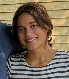 Young woman with short brown hair smiling, wearing a black and white striped shirt, standing outdoors in front of a wooden wall.