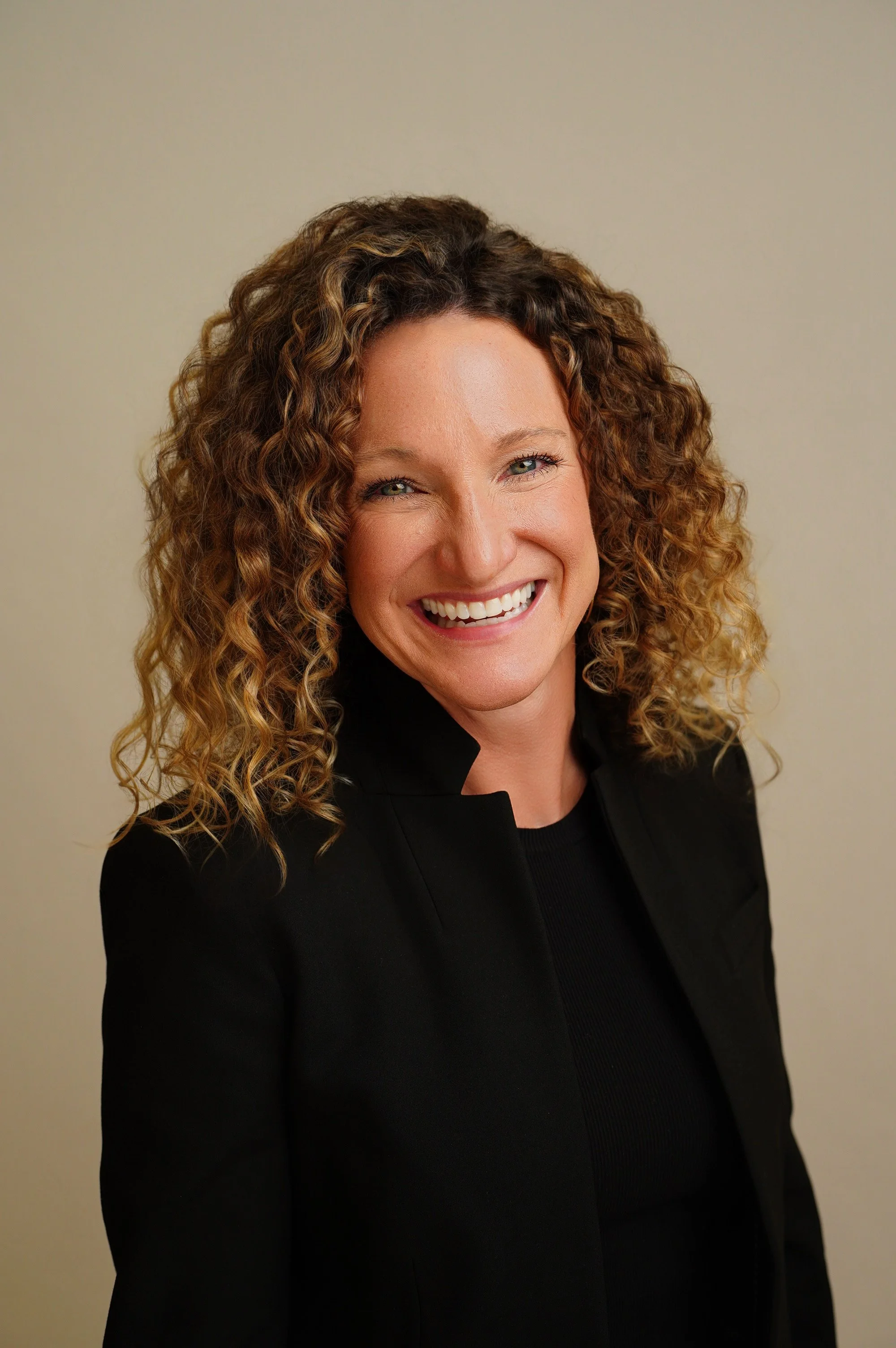 A woman with curly brown hair smiling and wearing a black jacket against a neutral background.