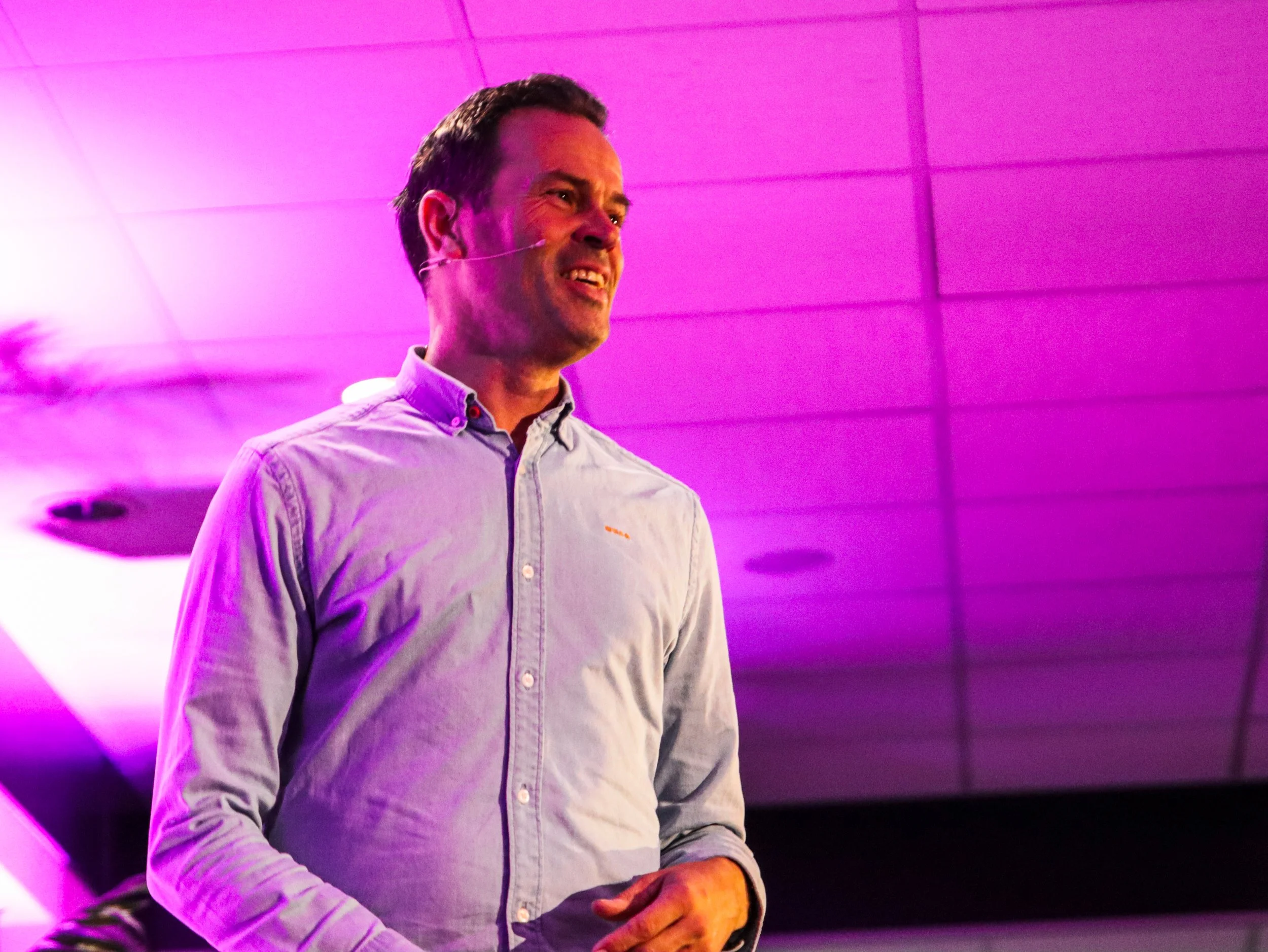 Man in een lichtblauw overhemd, sprekend tijdens een presentatie onder roze en paarse verlichting.