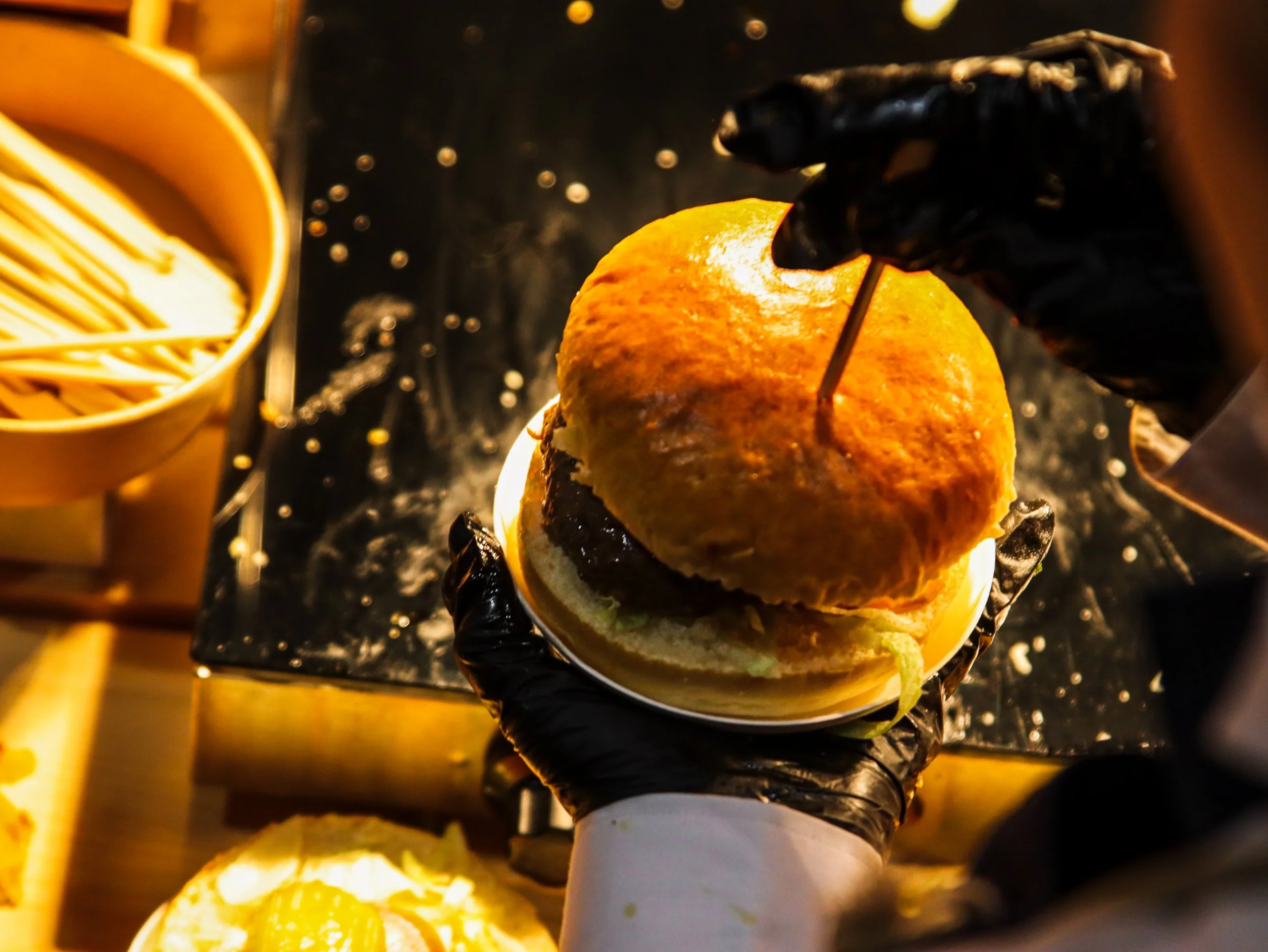 Persoon die een hamburger in een keuken bereidt, met een handschoen die een vork vasthoudt die in de hamburger is gestoken.