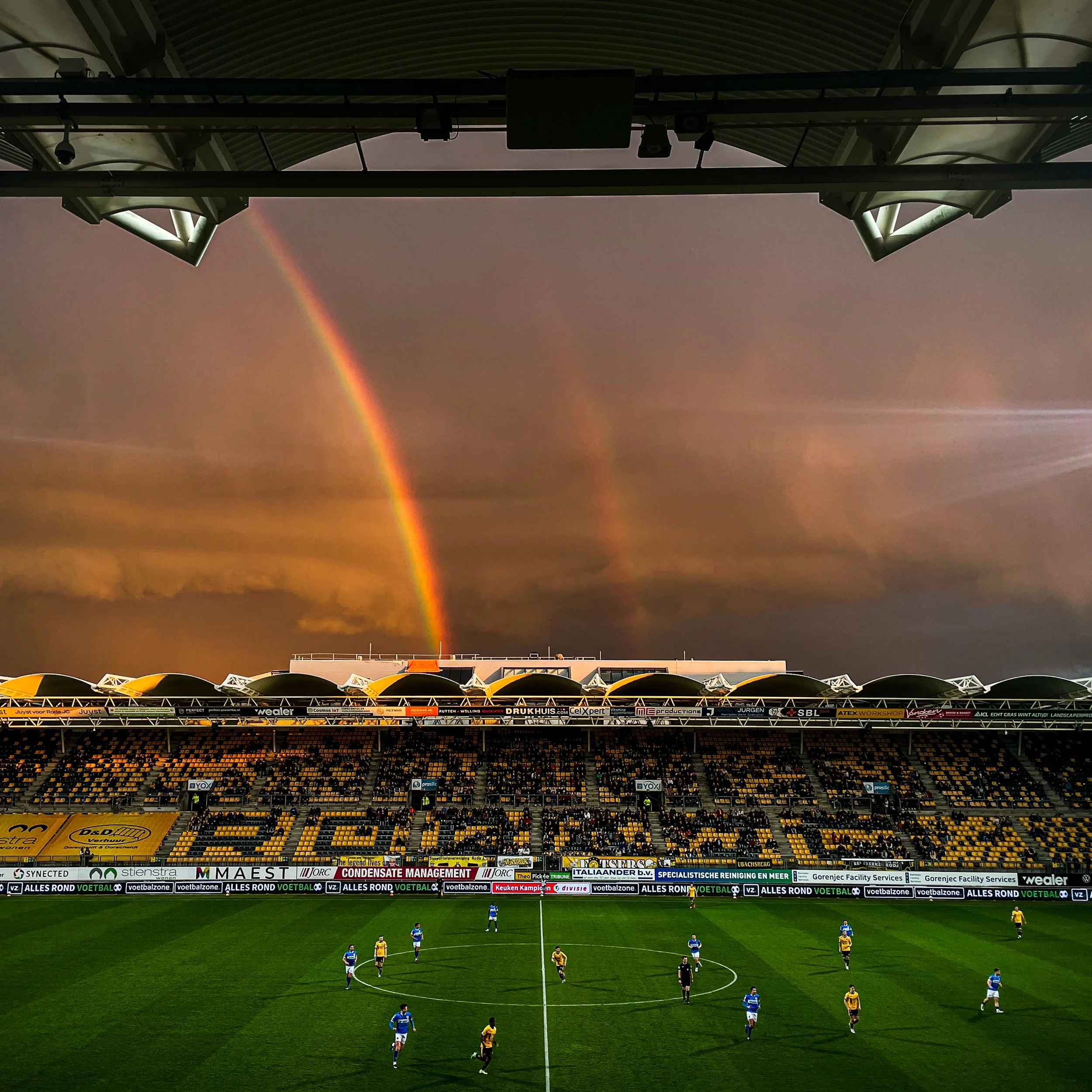 Een voetbalwedstrijd wordt gespeeld op een stadion met een regenboog en een dubbele regenboog in de lucht op de achtergrond.