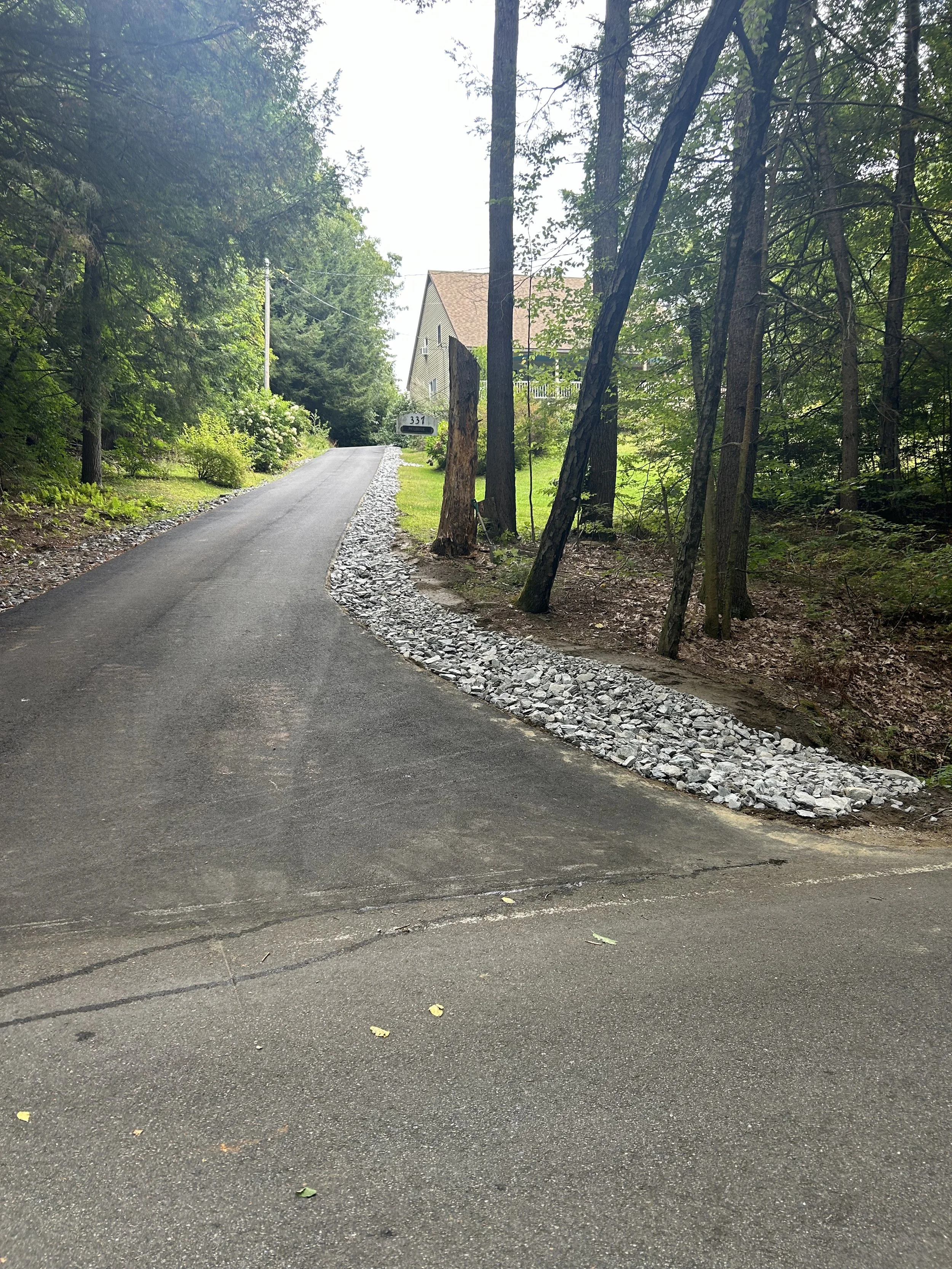 A paved driveway leading up to a house in a wooded area, with trees lining both sides and a rocky border along the edge of the driveway.