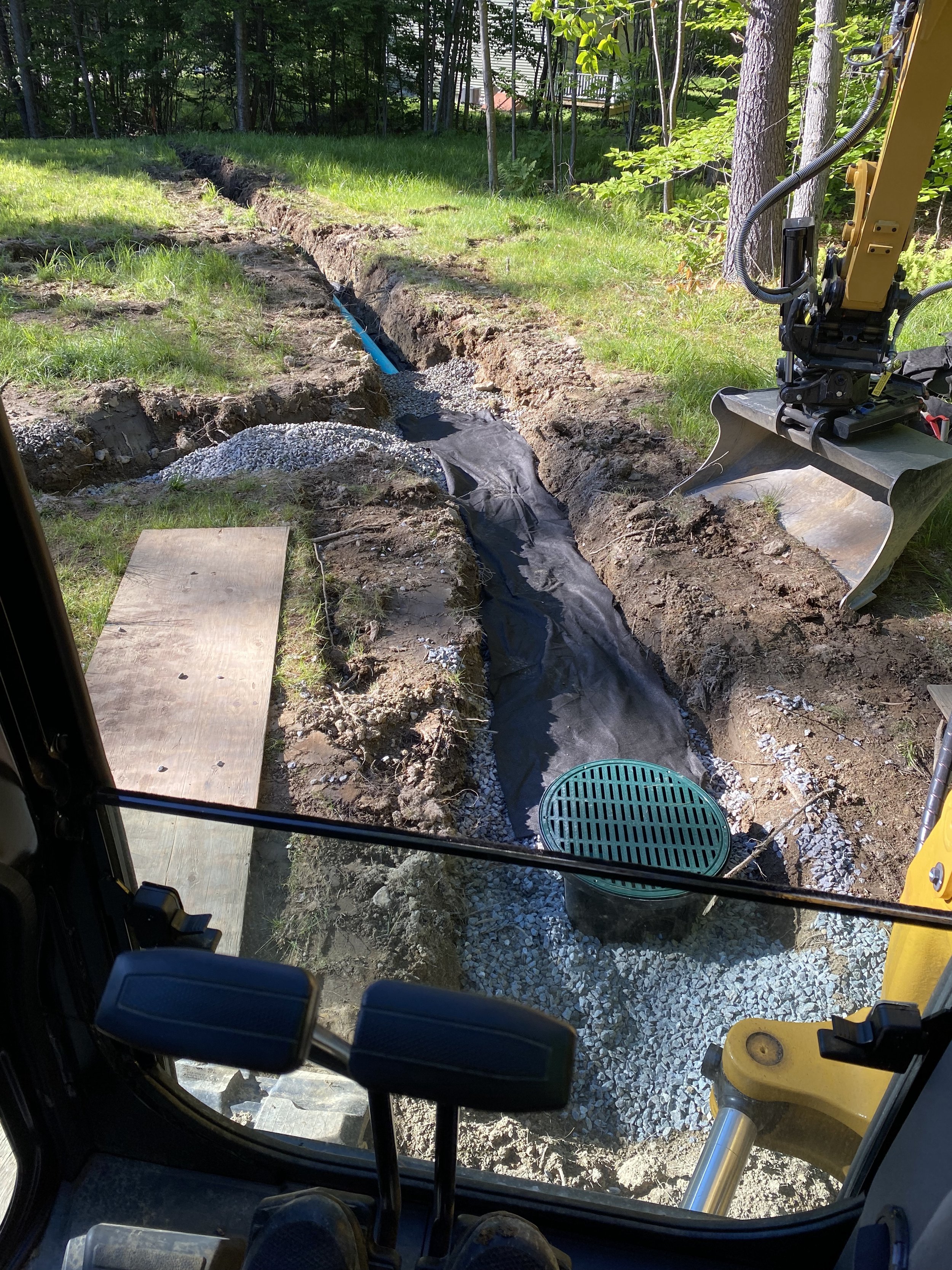 View from inside a construction vehicle showing a trench with black waterproofing fabric, gravel, and a blue pipe, with construction equipment nearby, in a grassy outdoor area surrounded by trees.