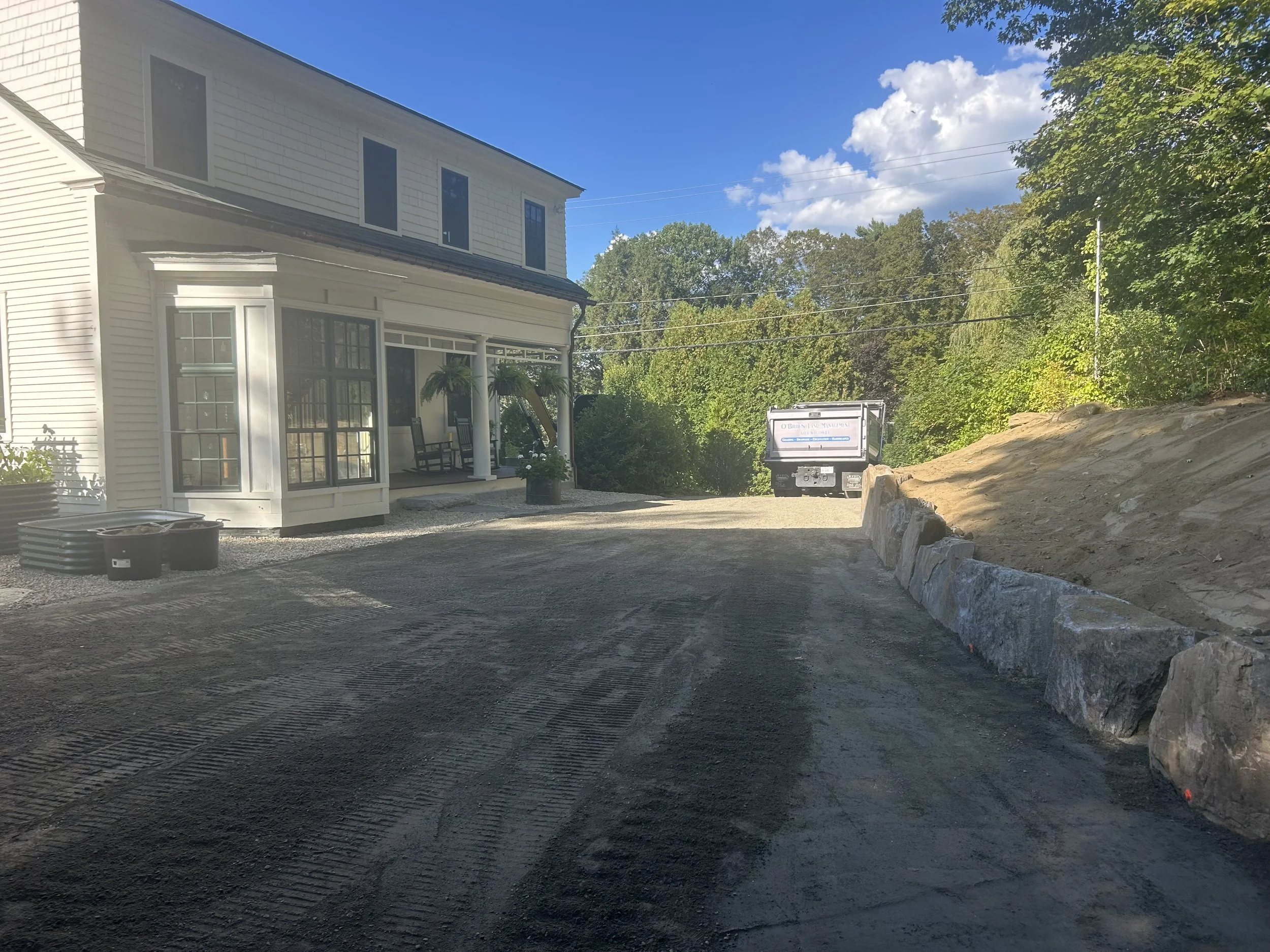A residential construction site with a new blacktop driveway, a white house with a porch, and a dump truck in the background. There are large rocks along the right side of the driveway and green trees on the right side.