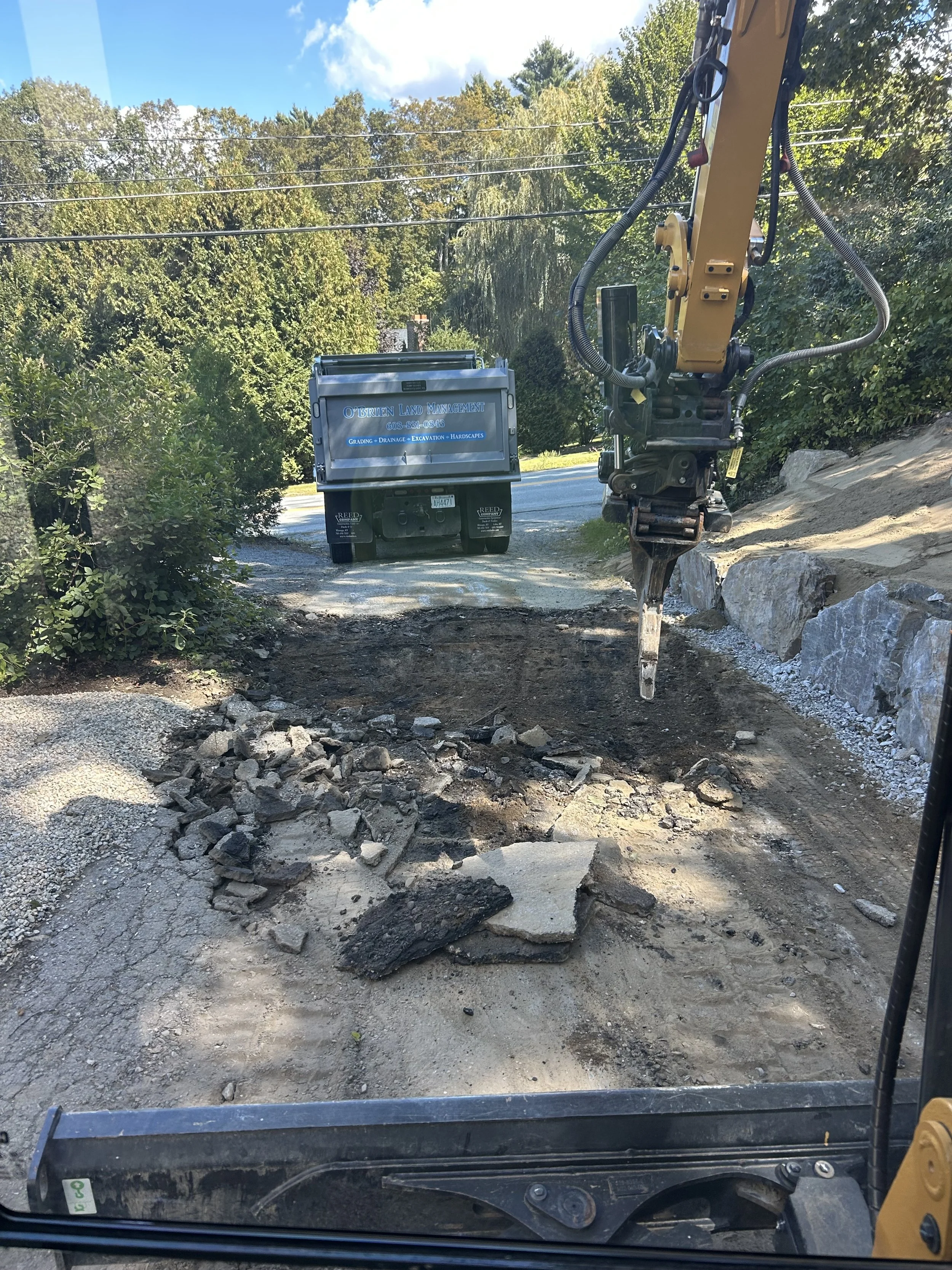 Construction site showing a small excavator removing rocks and soil from a pathway, with a large blue container in the background and trees on both sides.