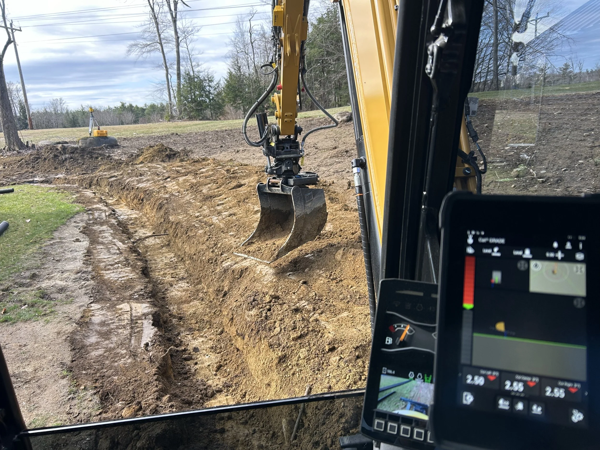View from inside a construction vehicle showing an excavator digging a trench on a grassy area with trees and cloudy sky in the background.